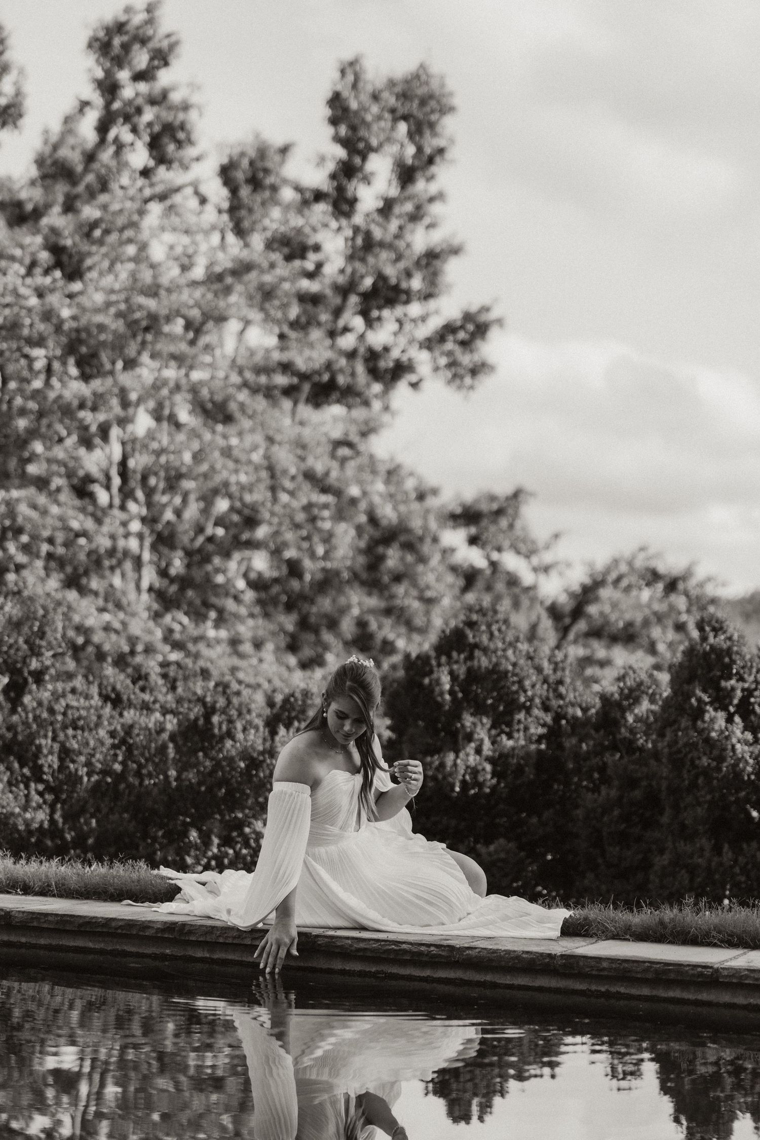 Black and white photo of a bride in a white gown sitting by a reflective pool surrounded by lush trees.