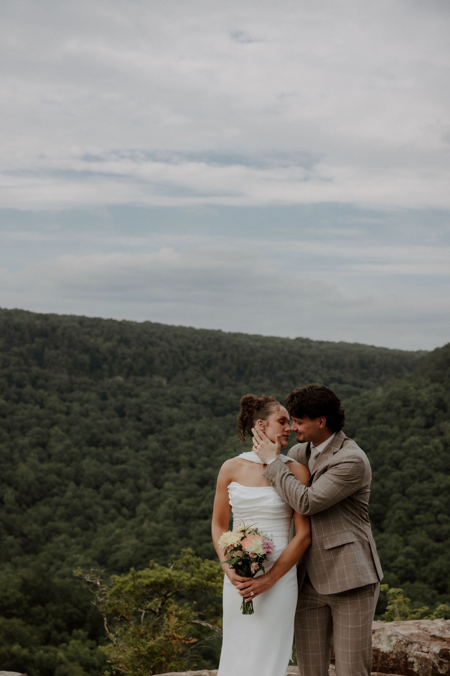 Bride and groom share a romantic moment on a scenic overlook with lush green forested mountains in the background.