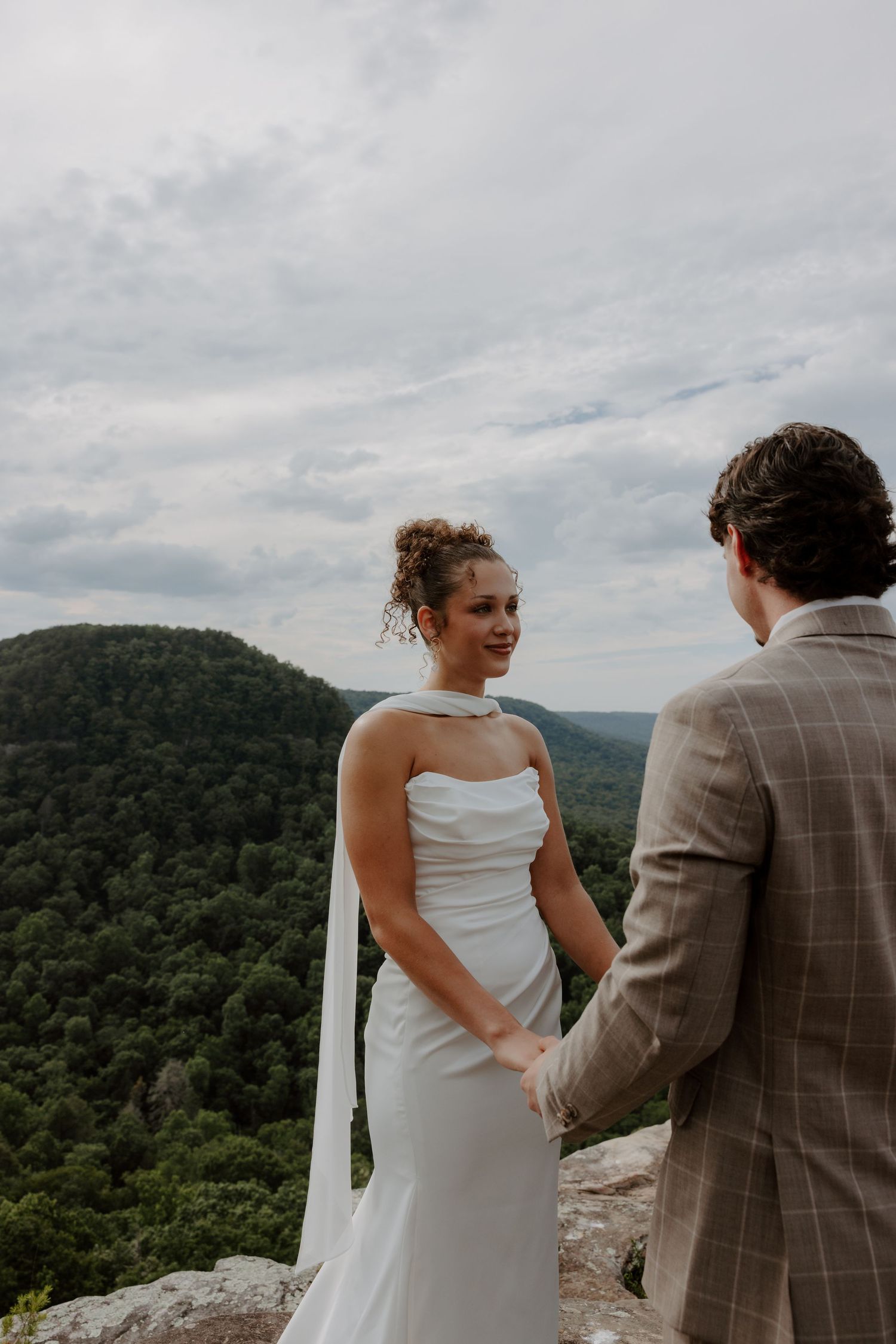 Bride in white gown holds hands with groom in plaid suit on rocky overlook with lush green mountains behind.