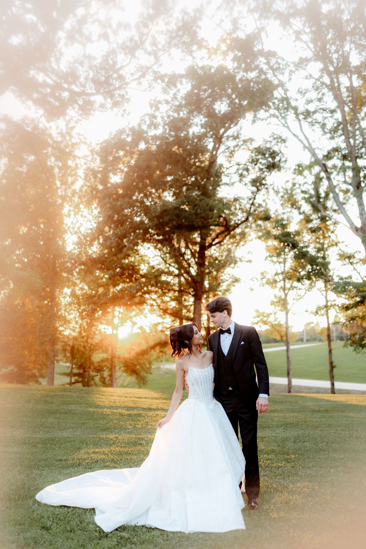 Bride and groom walking at sunset in a lush green park, bride in flowing white gown.