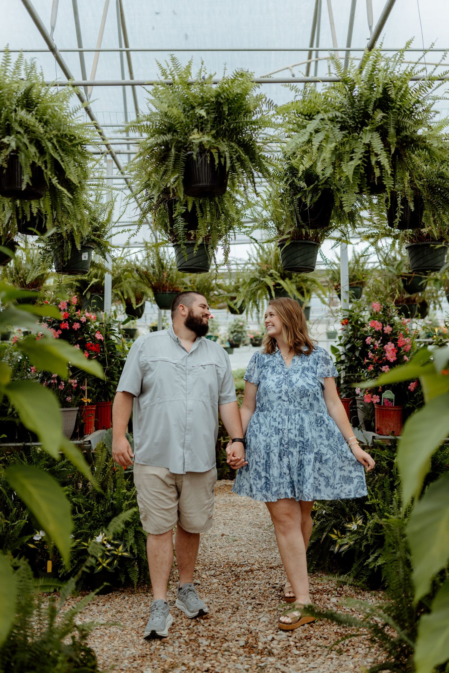 Couple holding hands and walking through a lush greenhouse filled with hanging ferns and colorful flowers.