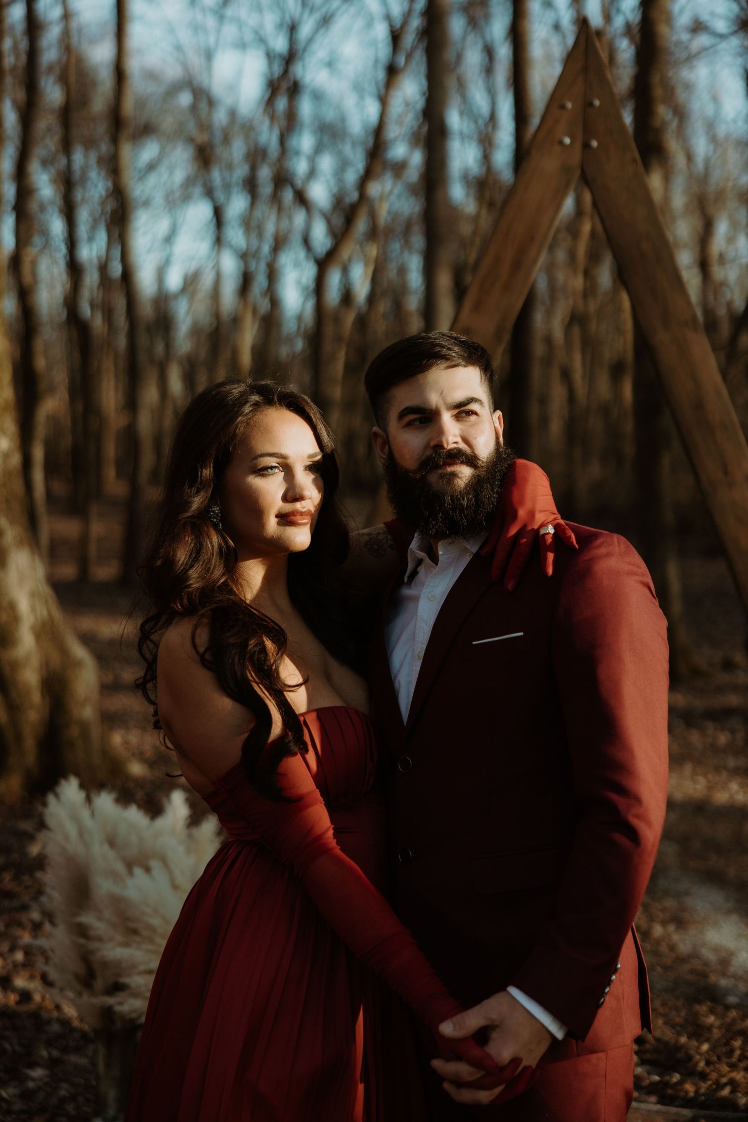 A couple in matching burgundy outfits pose romantically in a forest setting with a wooden arch backdrop.