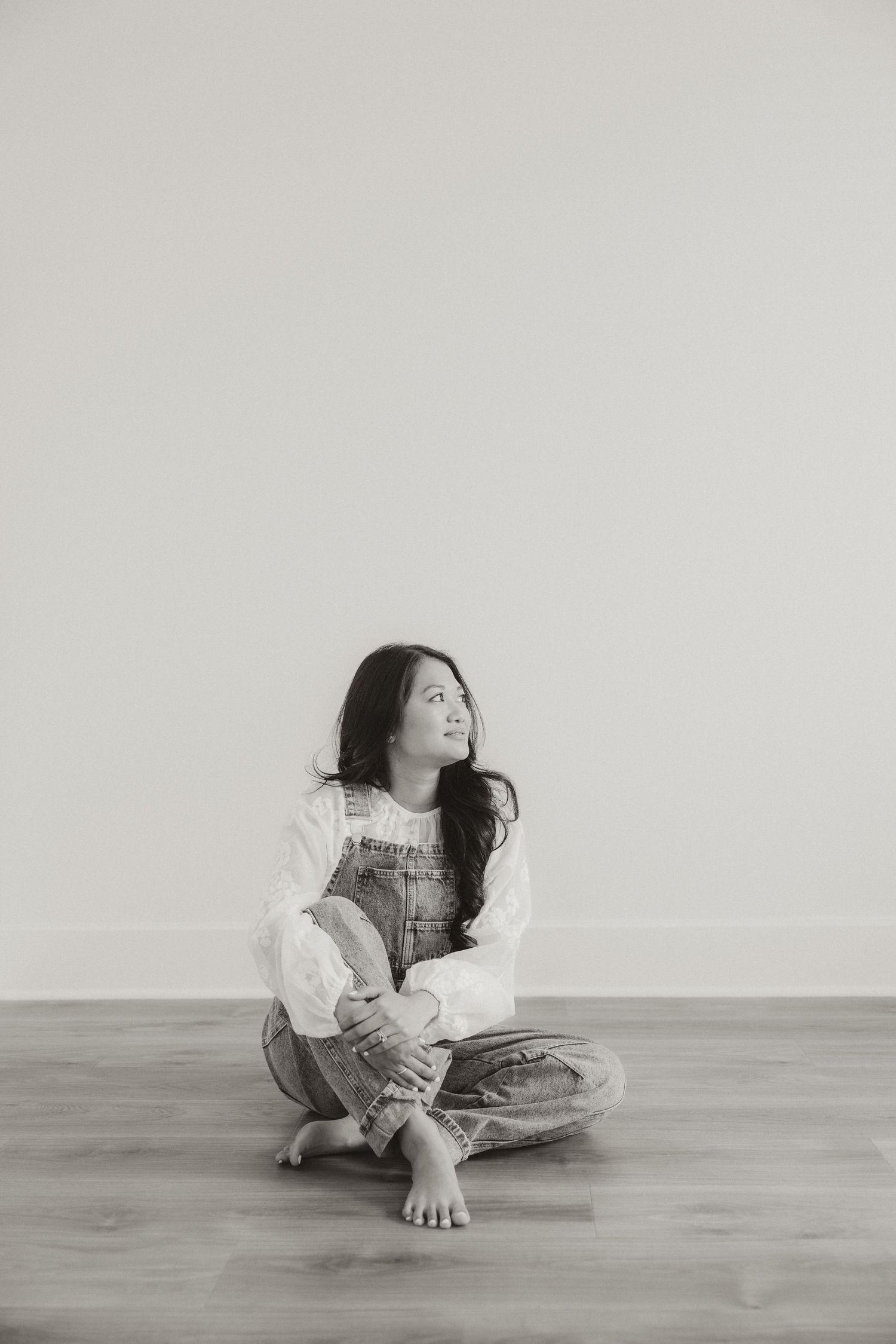 Black and white photo of a young woman sitting cross-legged on a wood floor wearing overalls, looking upward thoughtfully.