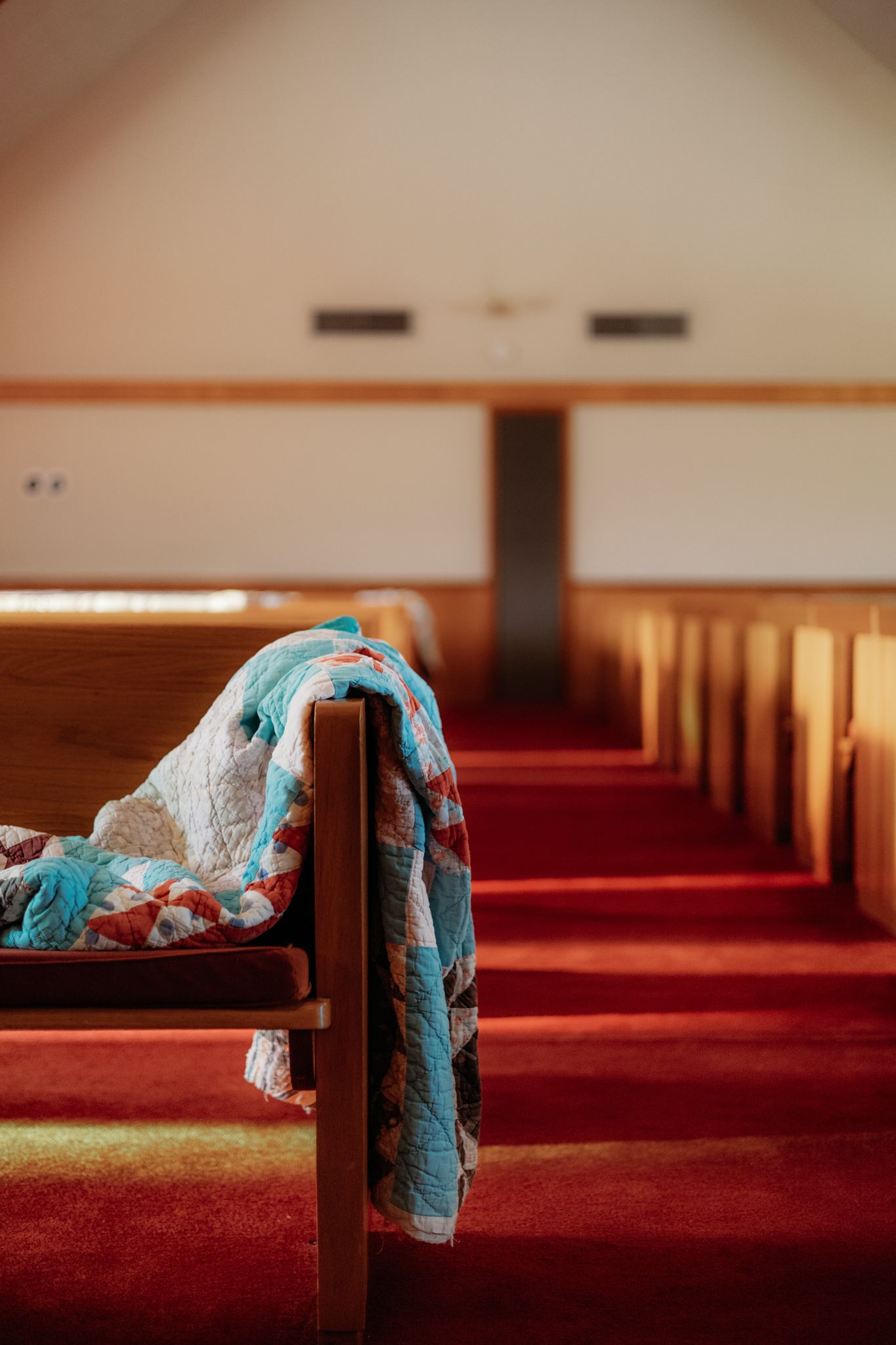 A blue and white quilt draped over a wooden church pew in an empty sanctuary with red carpeting.
