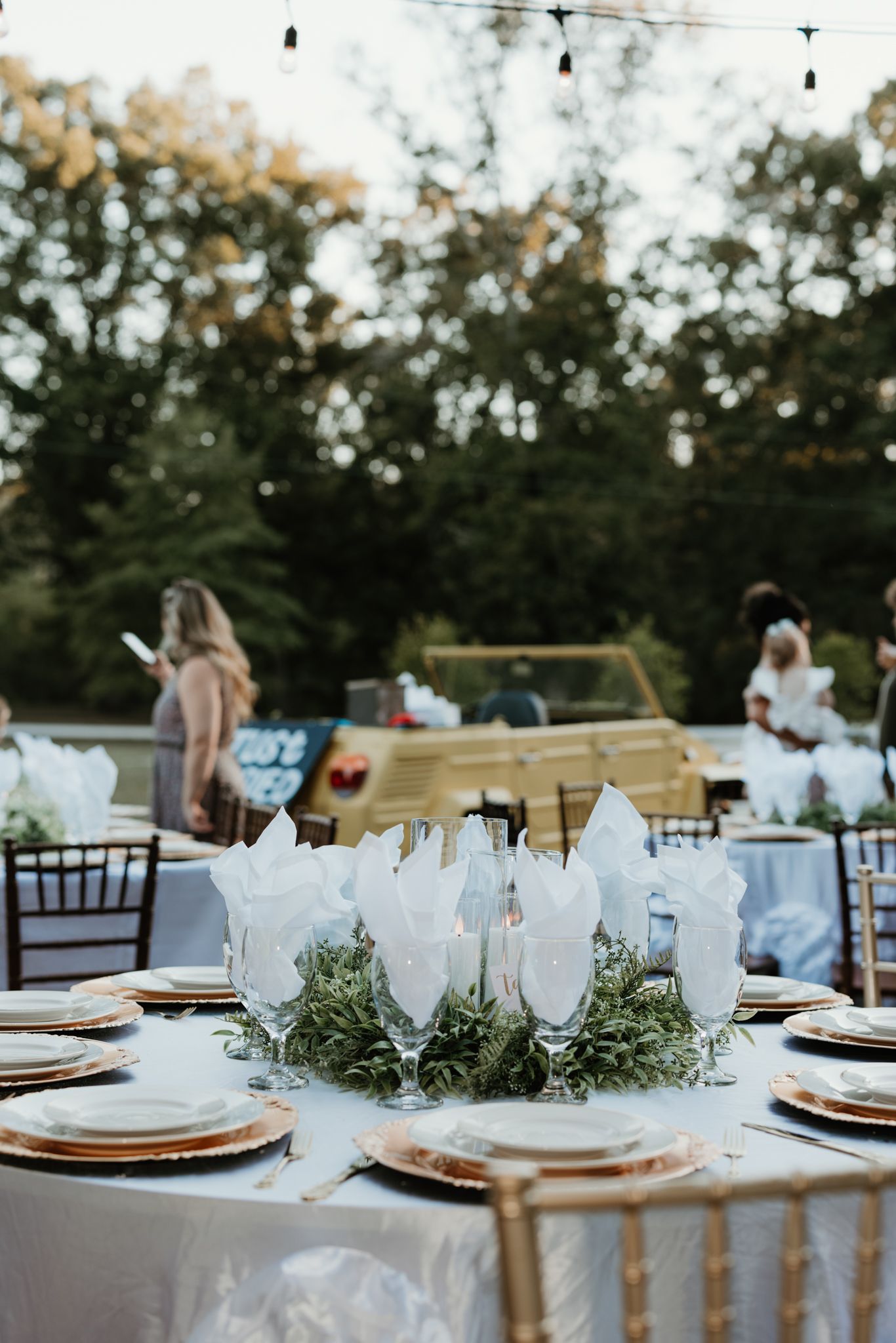 A wedding reception table set with gold chargers, white linens and crystal glassware amid string lights at an outdoor venue.