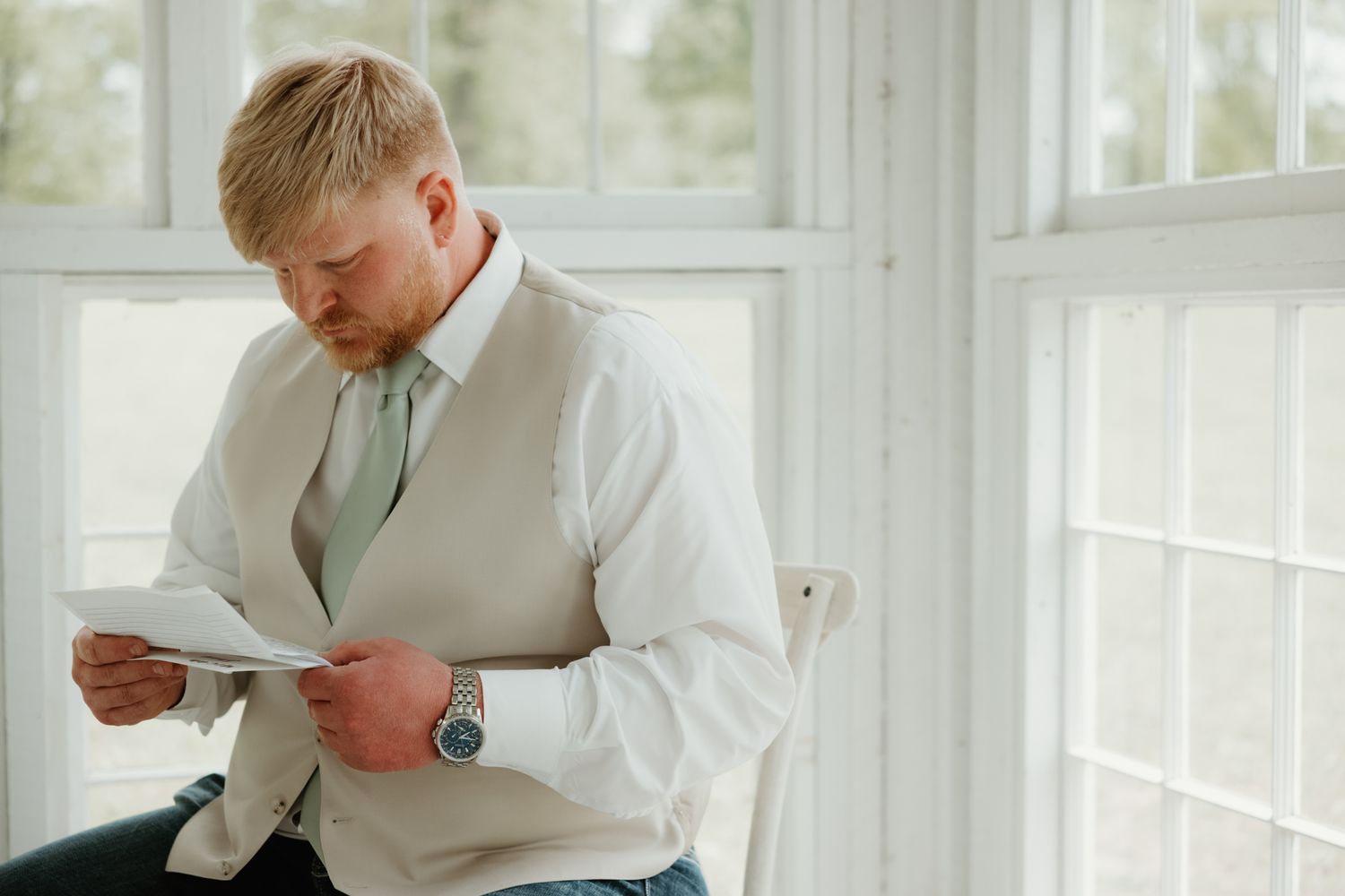 Someone wearing a cream vest and mint tie reads a document near bright sunlit windows.