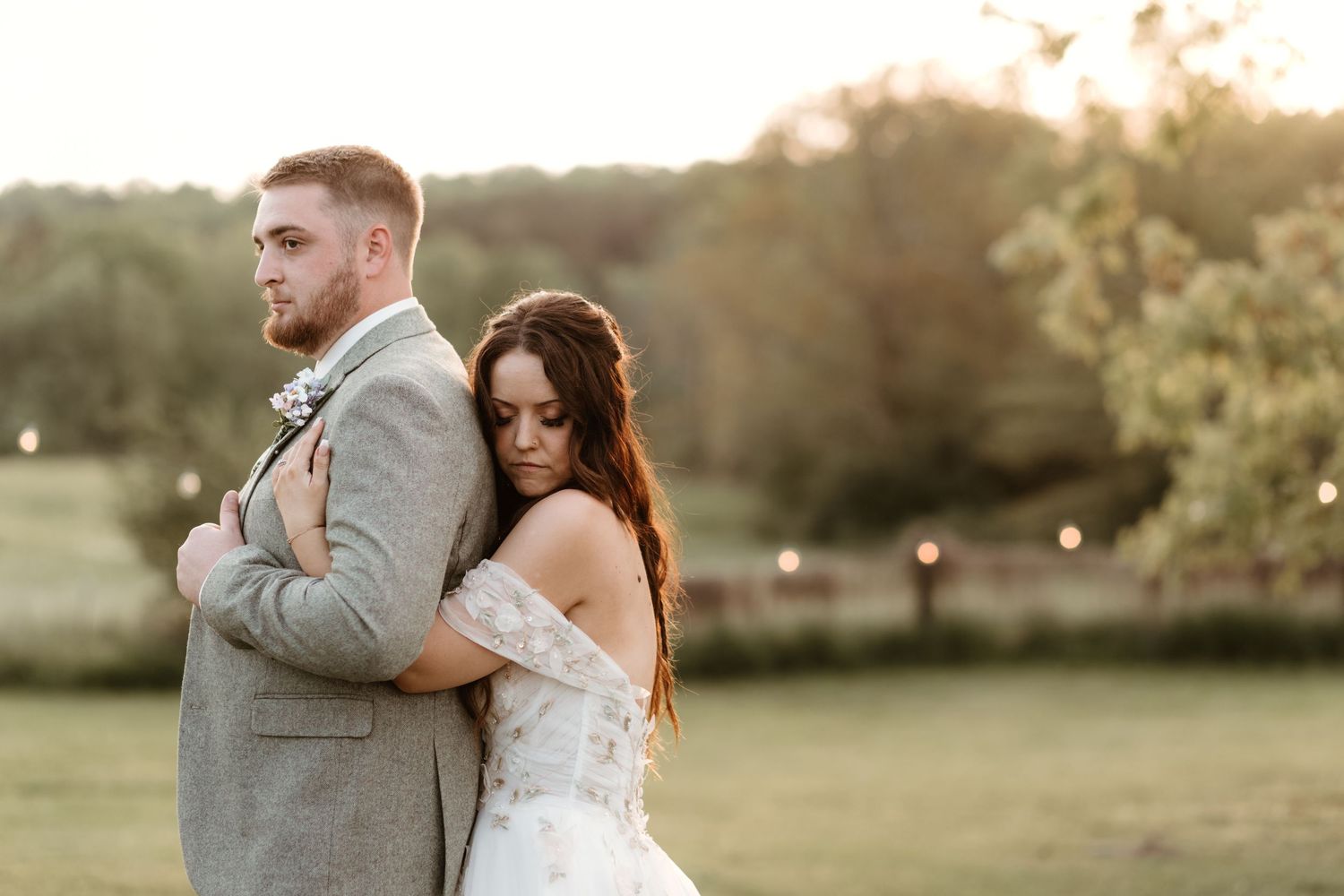 A bride and groom pose together in a grassy field during golden hour sunset.