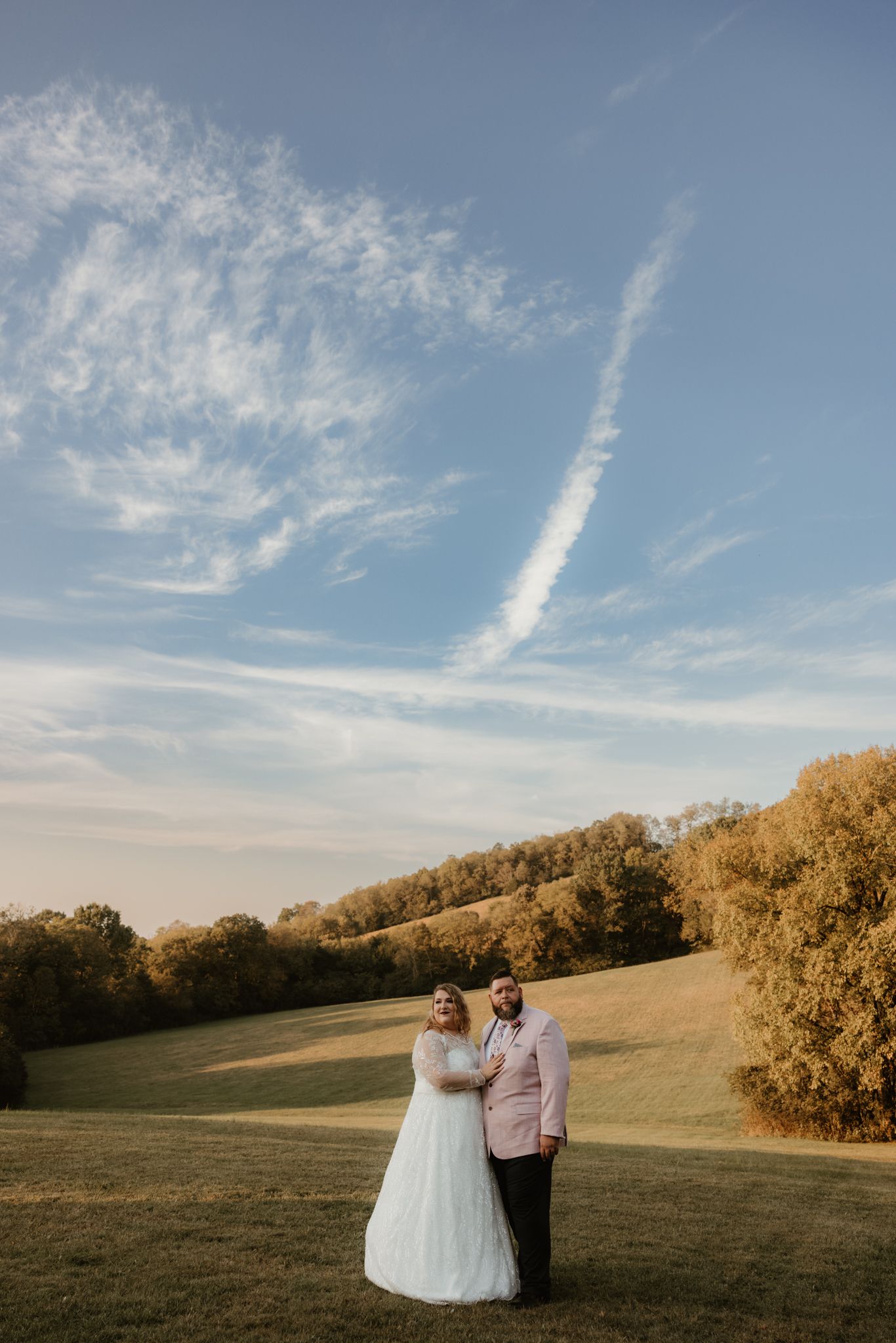 Couple in wedding attire posing in golden field beneath dramatic cloudy sky at sunset.