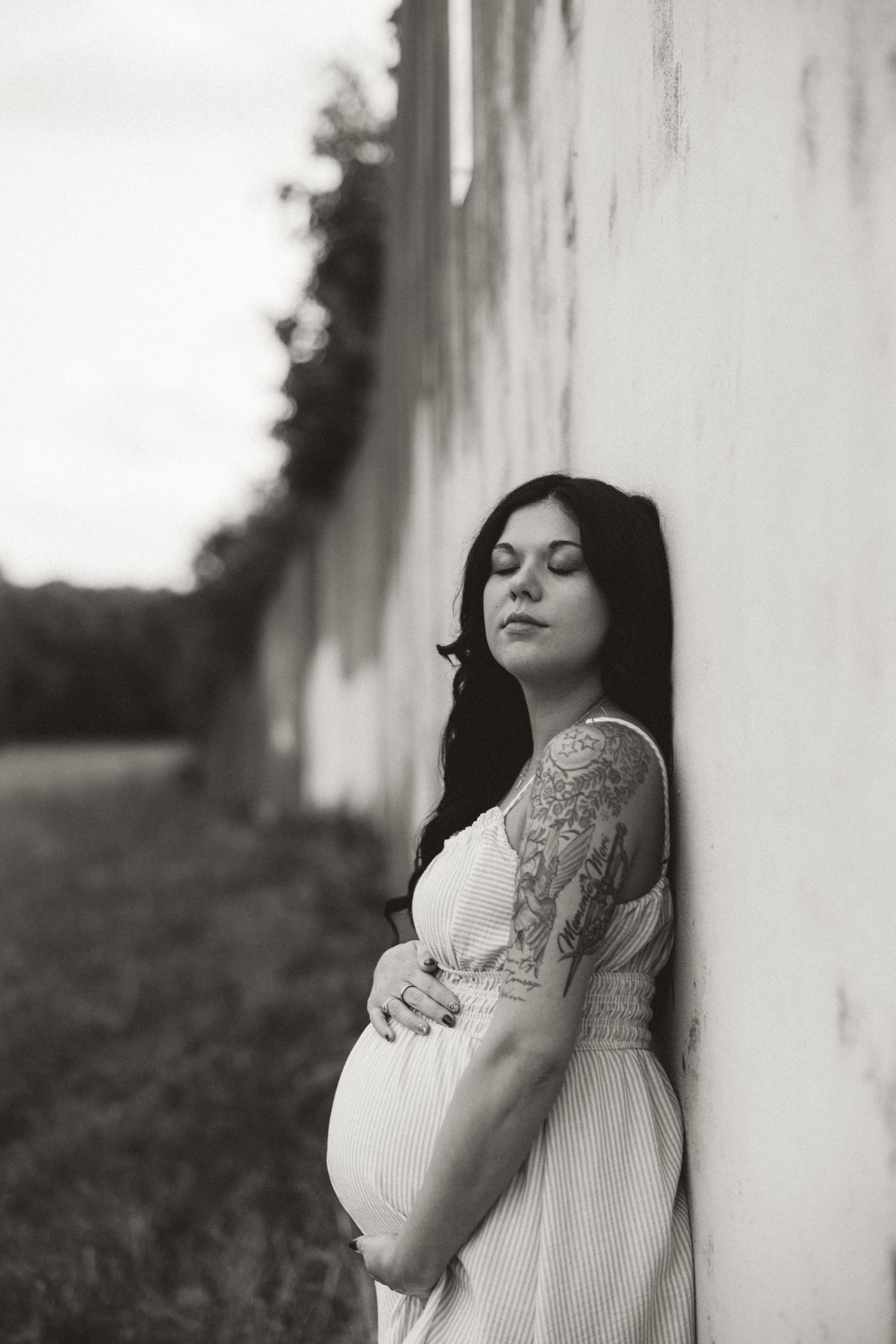 Black and white portrait of an expectant mother in a white dress leaning against a wall outdoors.