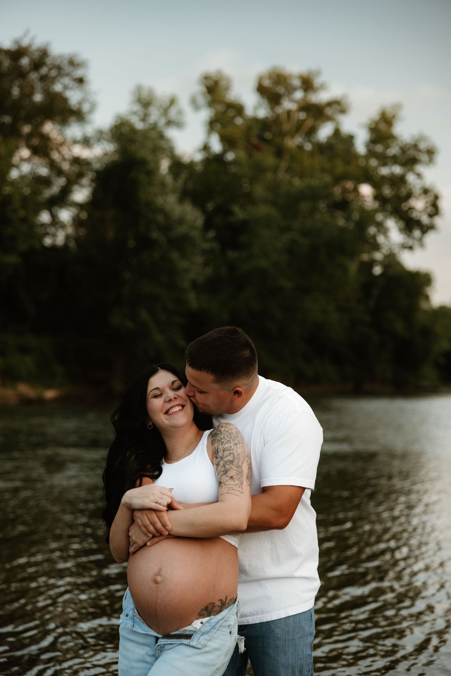 A couple shares a tender moment by the water during a maternity photoshoot at sunset.