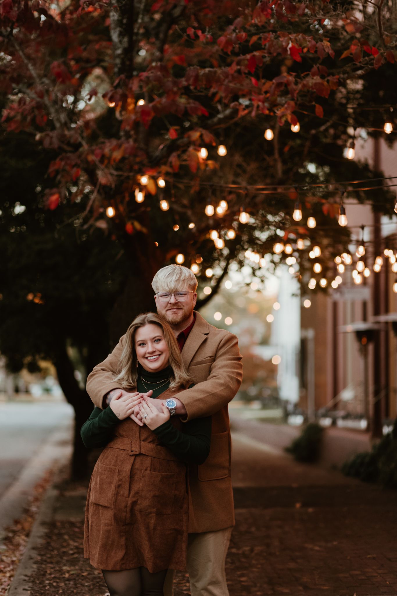 Two people embrace on an autumn evening street with twinkling string lights and red leaves overhead.