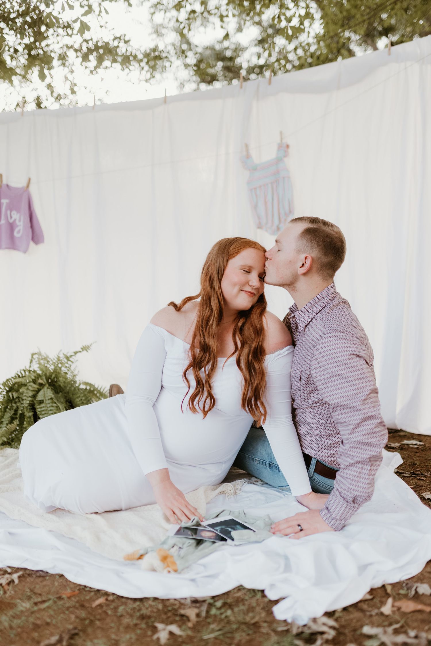 A romantic couple sits on a white blanket outdoors with a clothesline and greenery in the background.