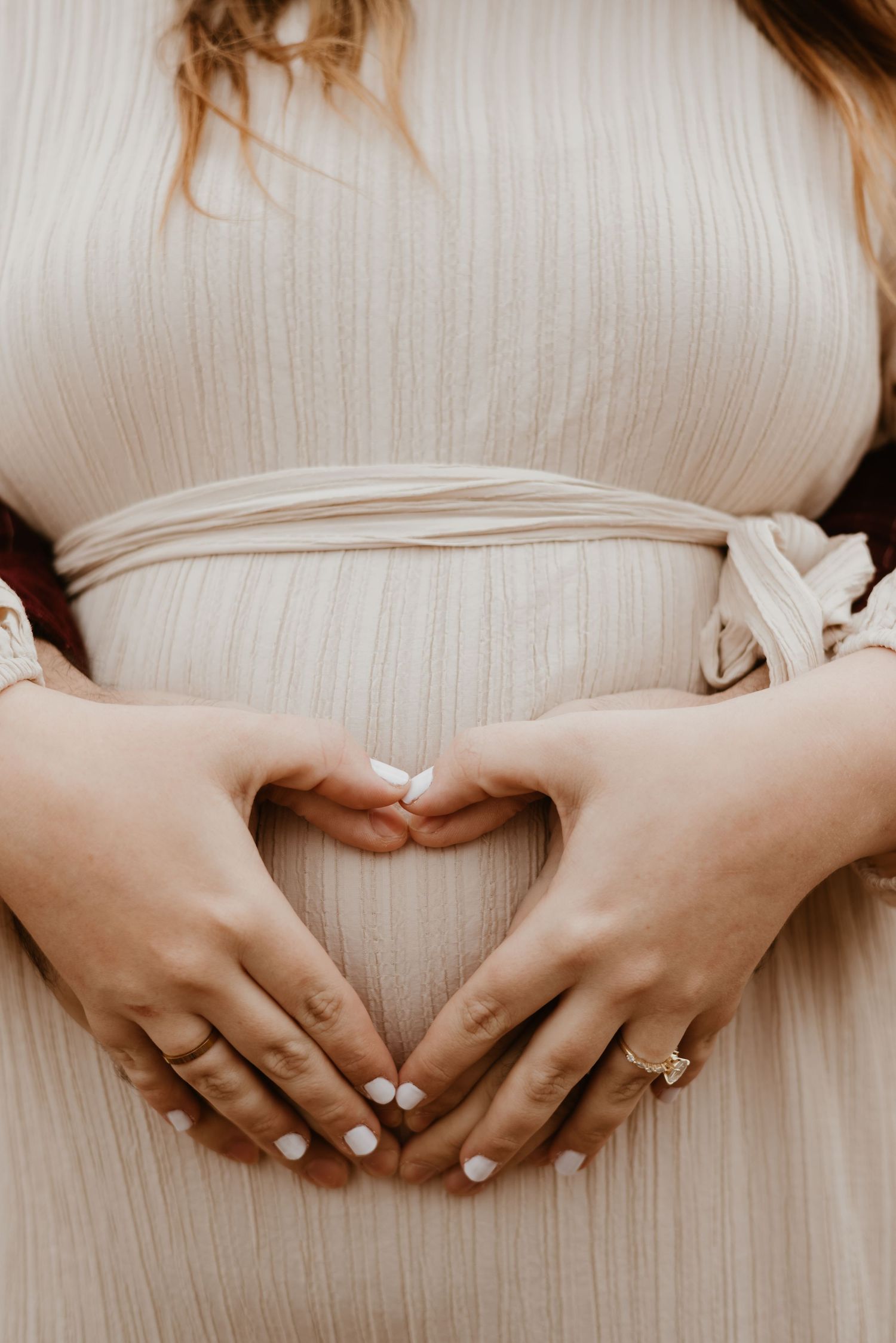 Close-up of hands forming a heart shape over a pregnant belly in a cream dress.