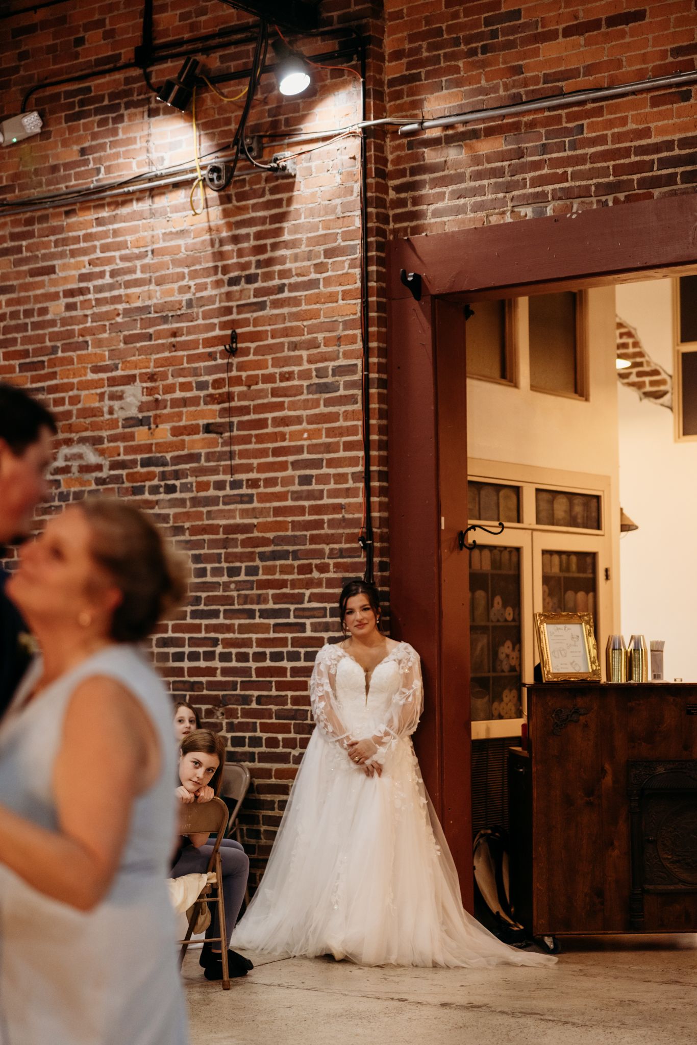 A bride in a long-sleeved lace gown stands in front of an exposed brick wall with industrial lighting.