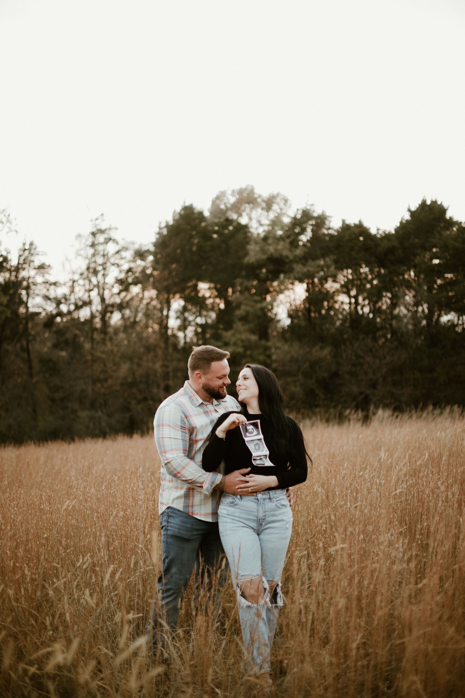 A couple stands together in a golden field of tall grass during sunset.