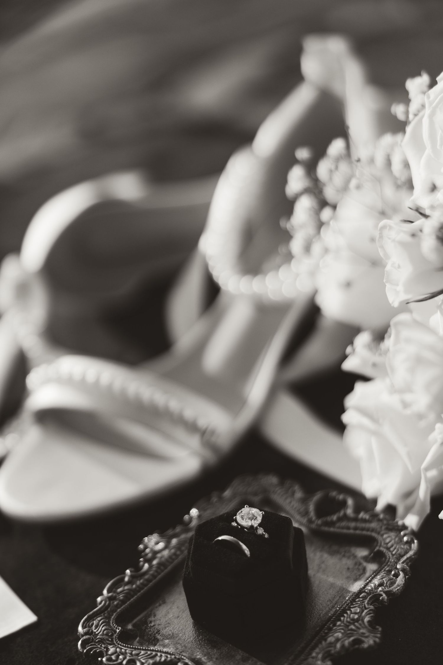 A diamond engagement ring on a black velvet cushion with shoes and white flowers in the background.