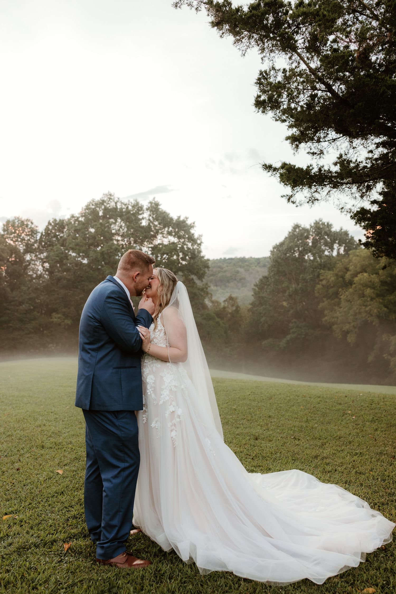 Romantic outdoor wedding photos taken in a misty field with flowing white dress and navy suit at sunset.
