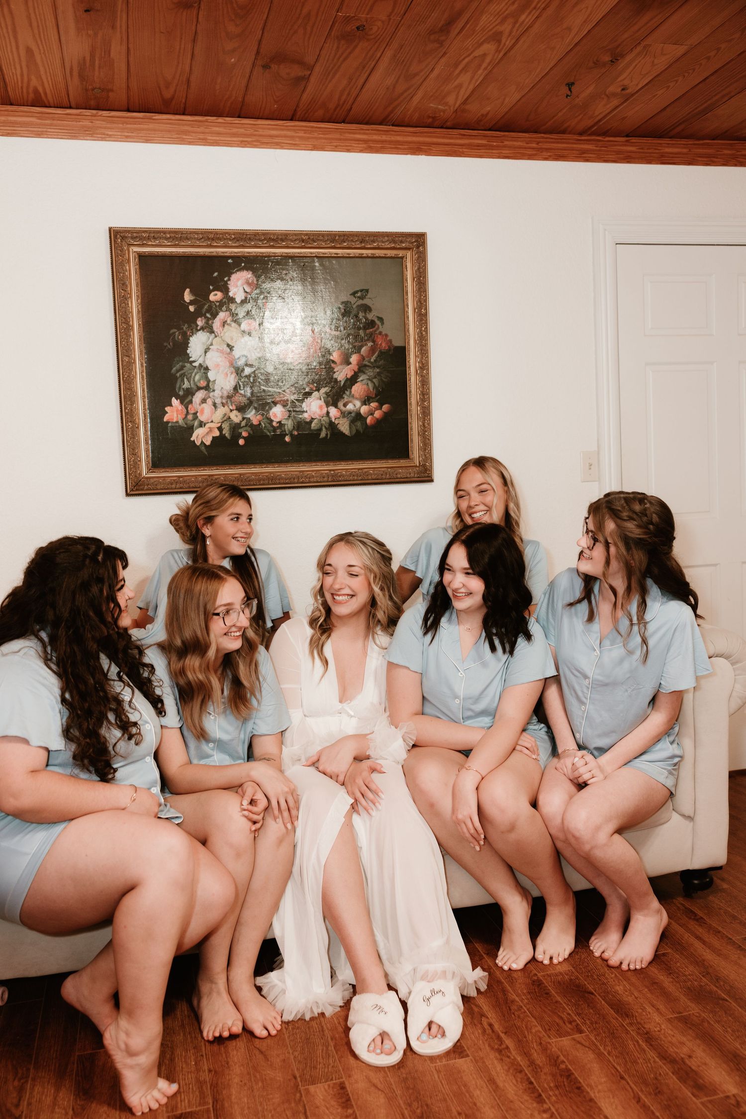 A group in matching blue shirts and white socks sit together on a hardwood floor against a white wall with framed artwork.
