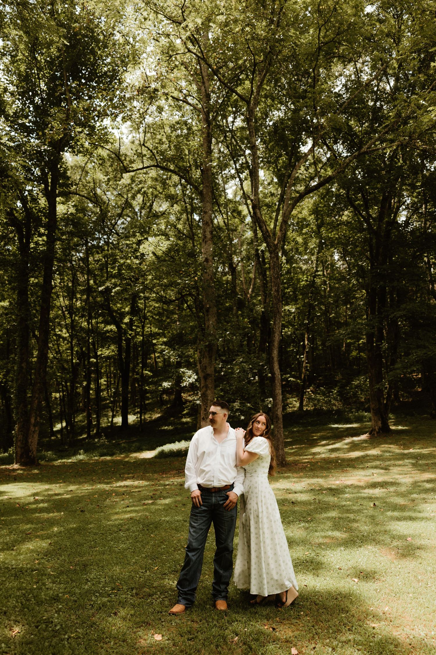 A couple stands together in a sunlit wooded area, surrounded by tall trees and dappled sunlight on a grassy lawn.