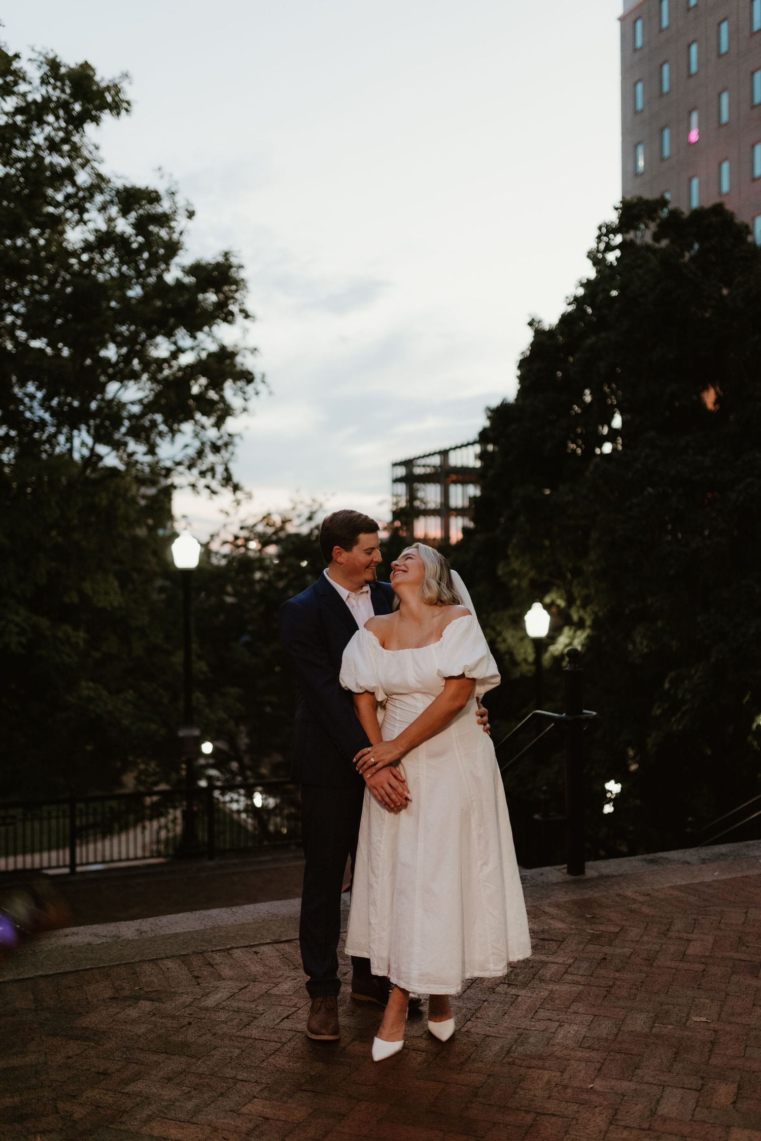 A newlywed couple shares an intimate dance moment together at dusk on a brick paved terrace with city buildings behind them.