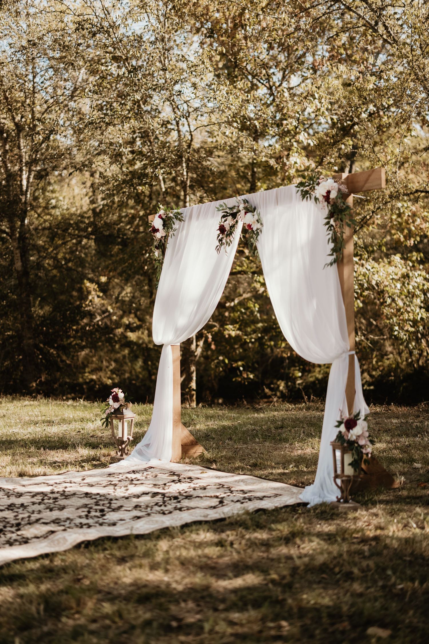 A sequence of wooden wedding arch frames with flowing white fabric in an outdoor garden setting at sunset.