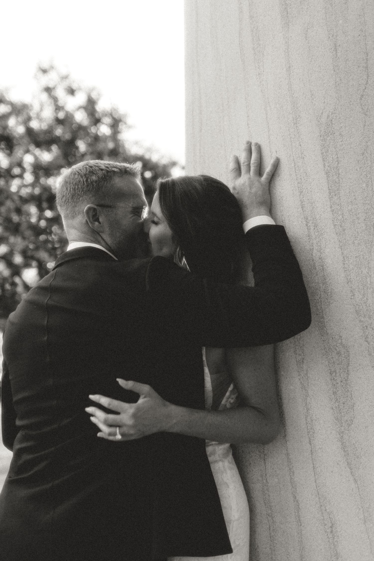 A black and white romantic photo of a couple embracing against a wall outdoors with trees in background.