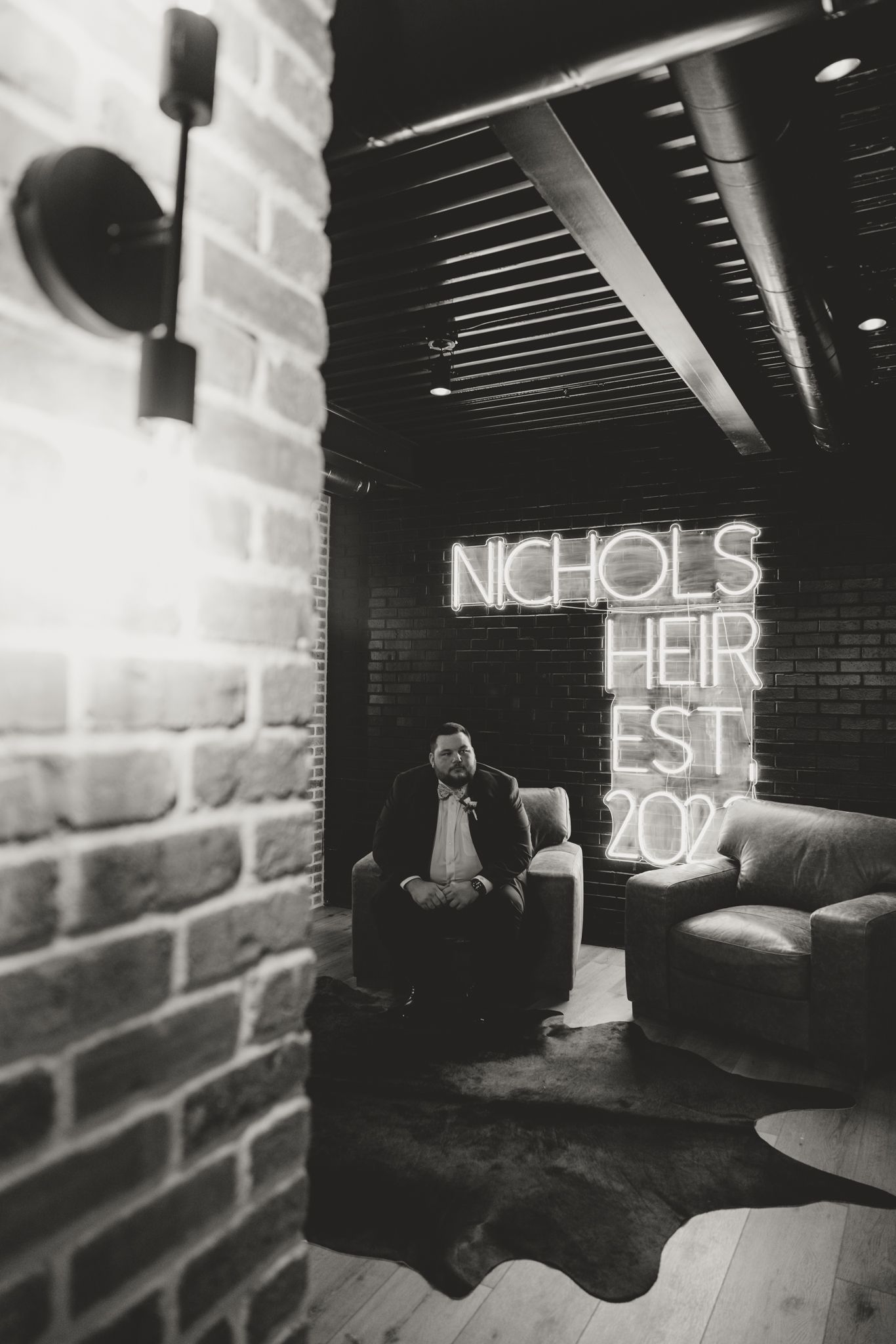 Interior space with rustic brick walls and wooden beams featuring illuminated 'NICHOLS EST' neon signage.