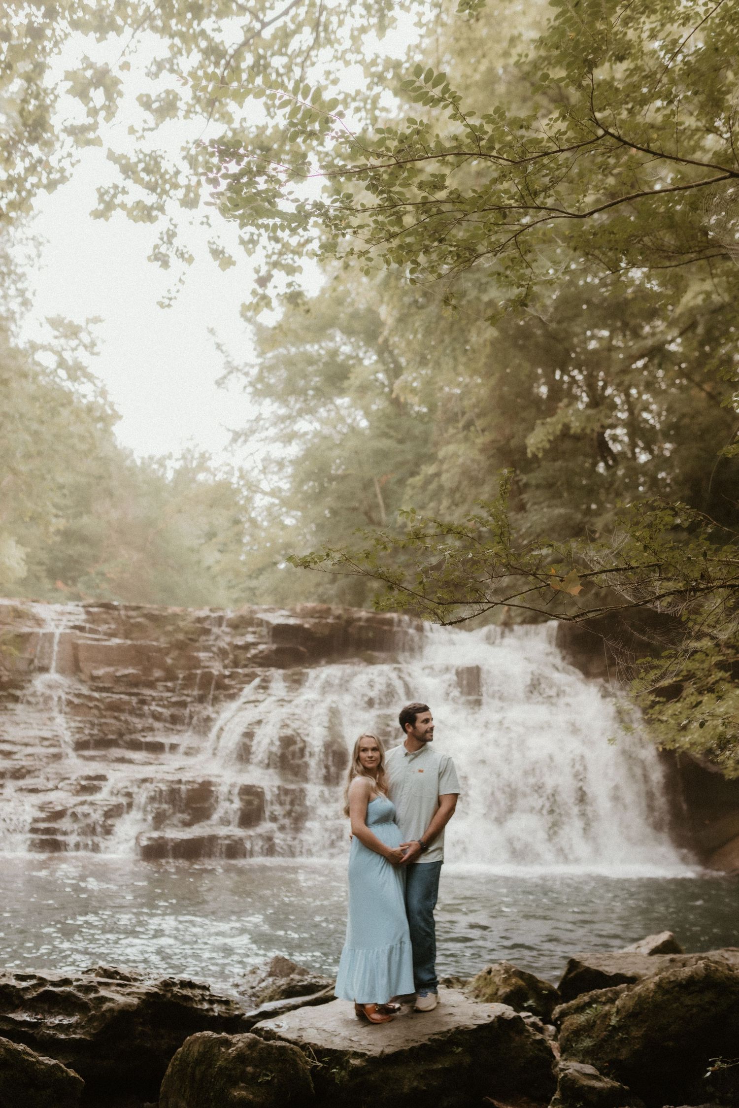 A couple poses on rocky outcrop in front of cascading waterfall during maternity session.