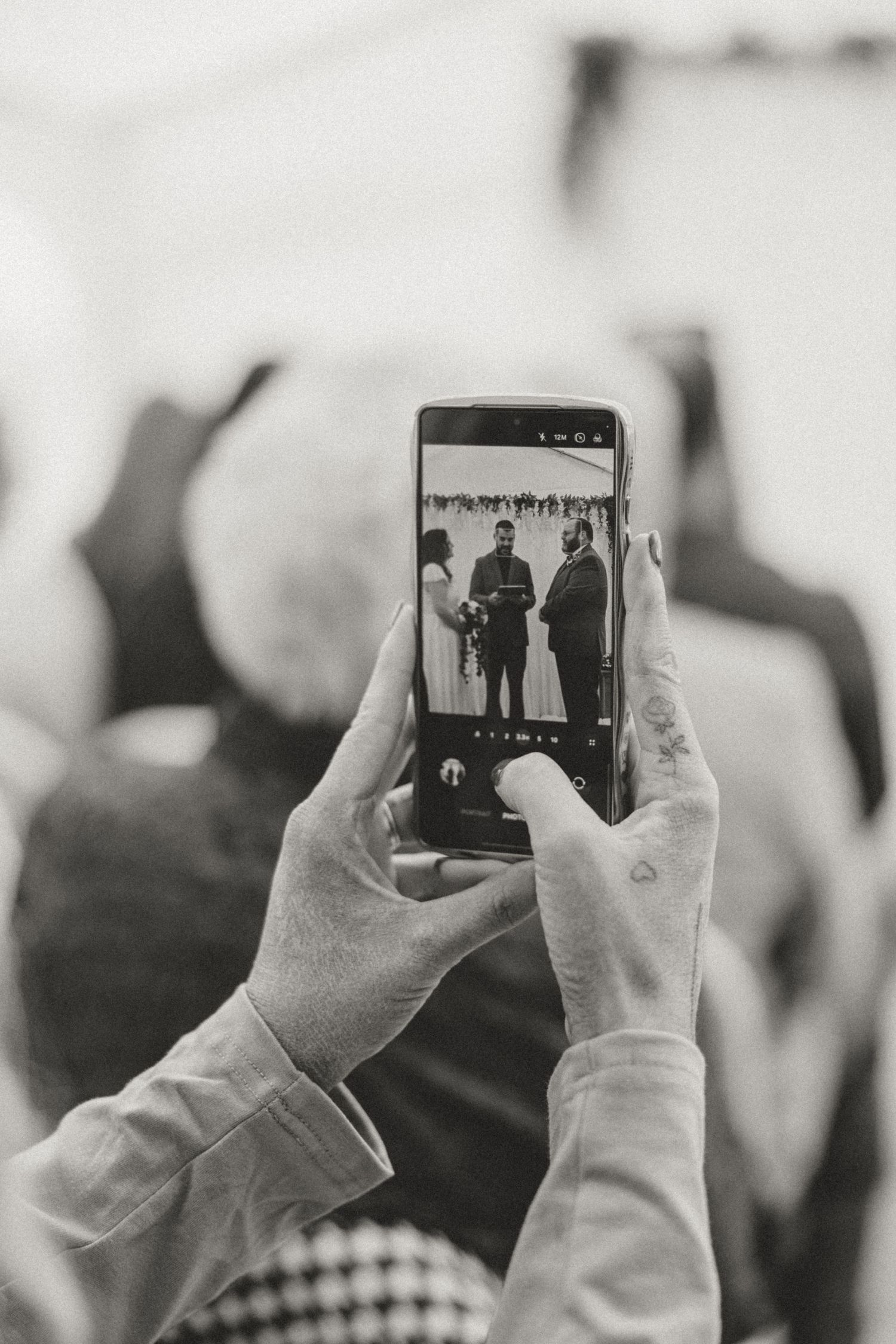 Black and white photo of hands holding a smartphone capturing a wedding ceremony with couple and officiant on screen.