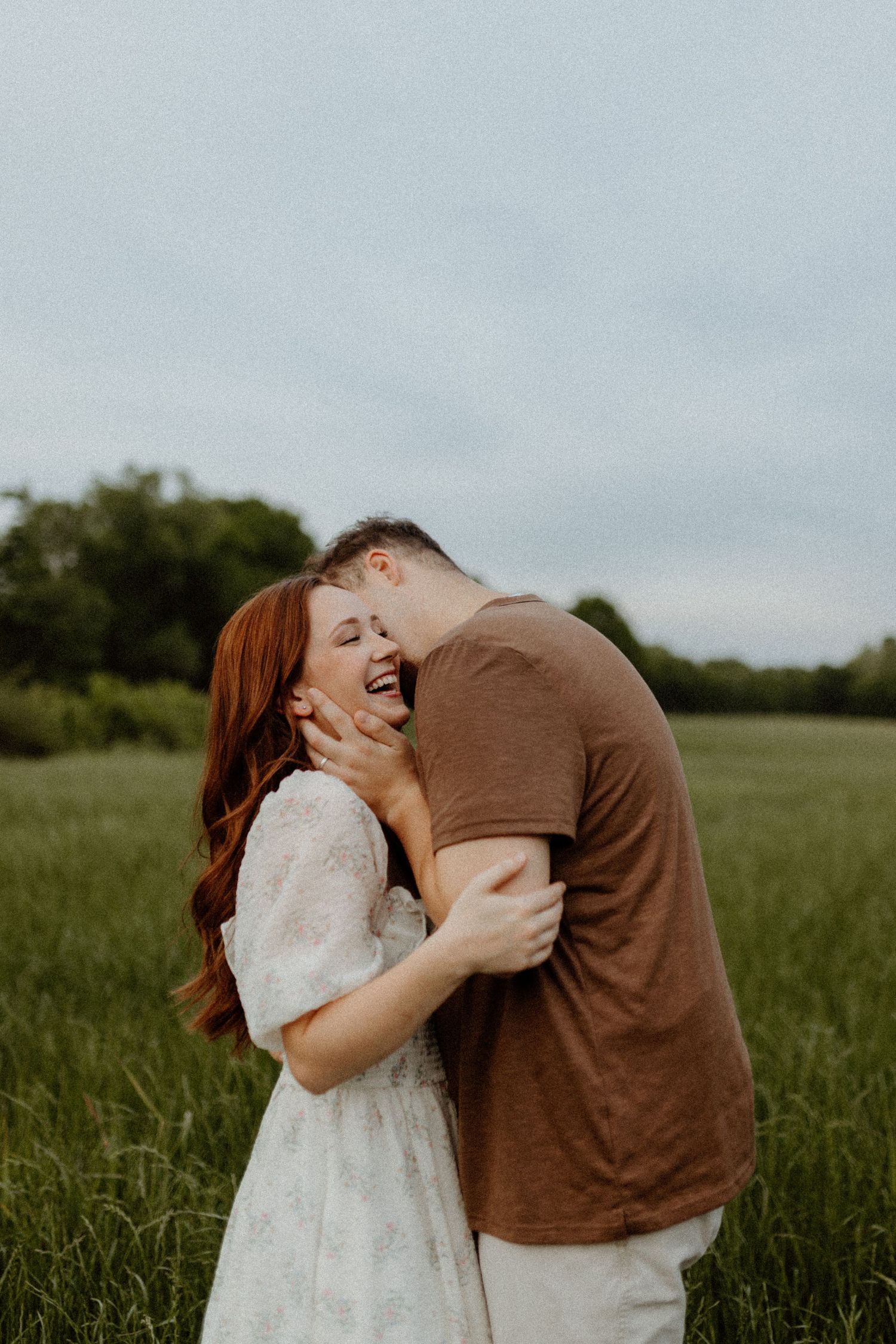 A joyful couple embraces and laughs together in a lush green field under a cloudy sky.