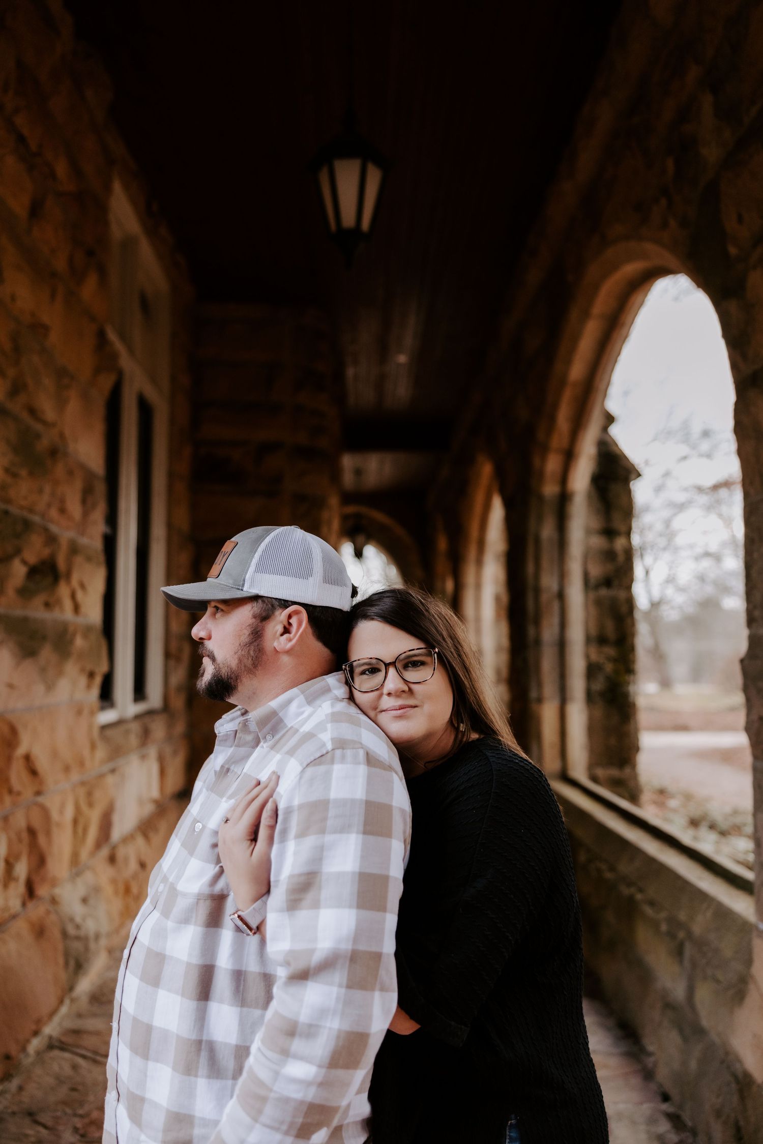Couple embracing in a stone archway corridor, woman in glasses leaning on mans back in plaid shirt and trucker hat.