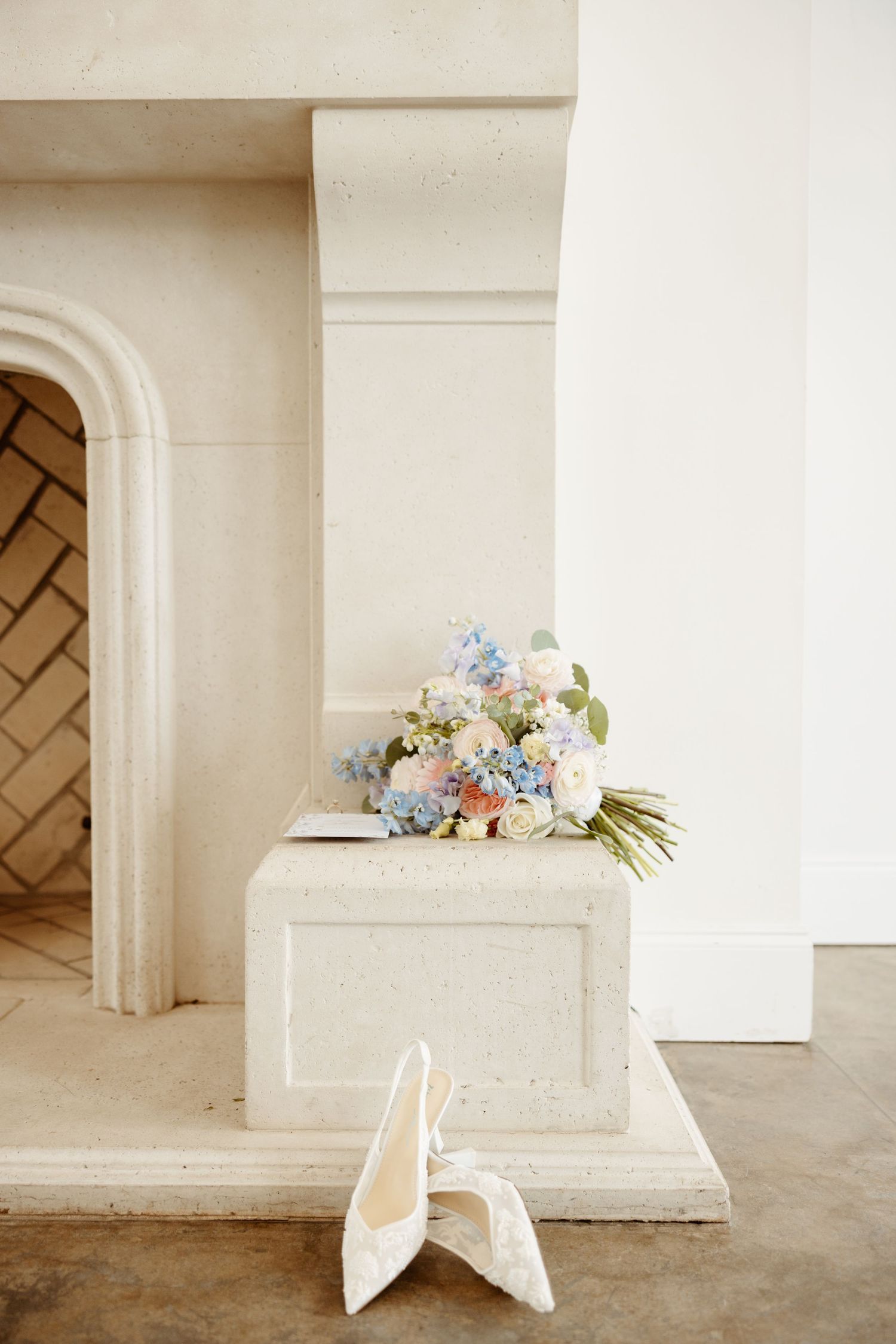 Elegant wedding bouquet with blue and pink flowers resting on a white fireplace mantel beside bridal heels.