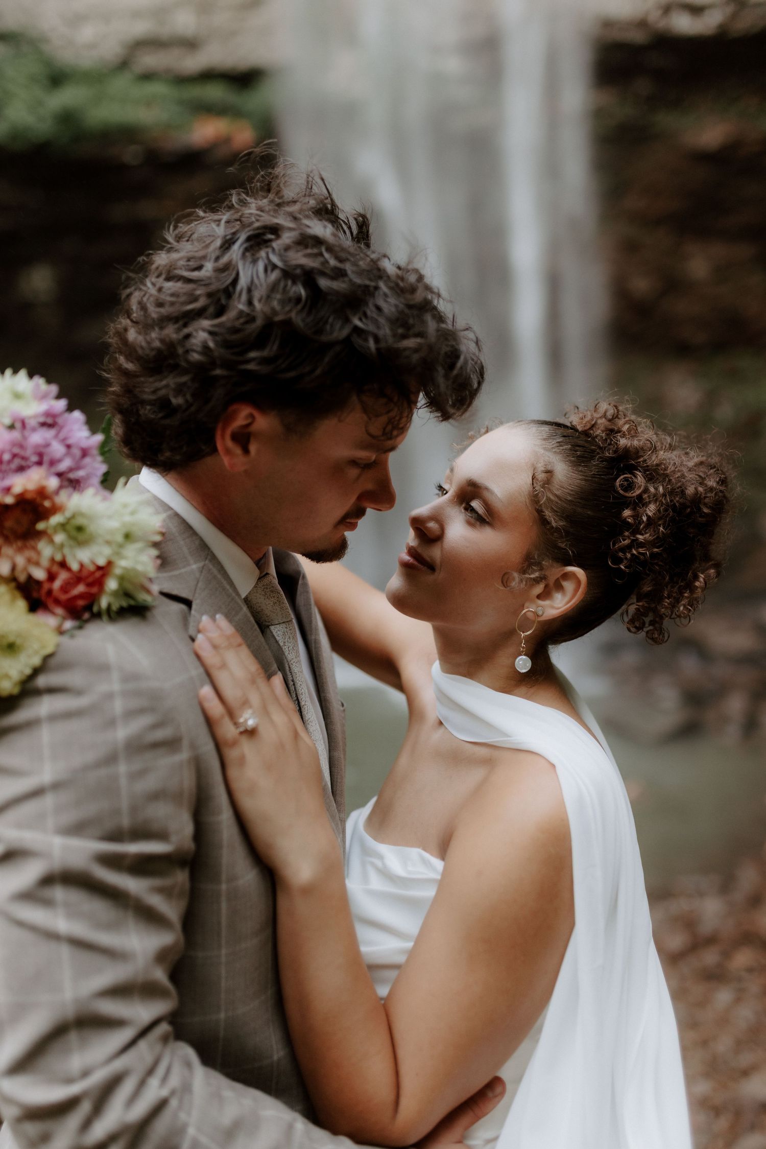 Bride and groom share an intimate moment in front of a waterfall, with bride holding colorful bouquet.