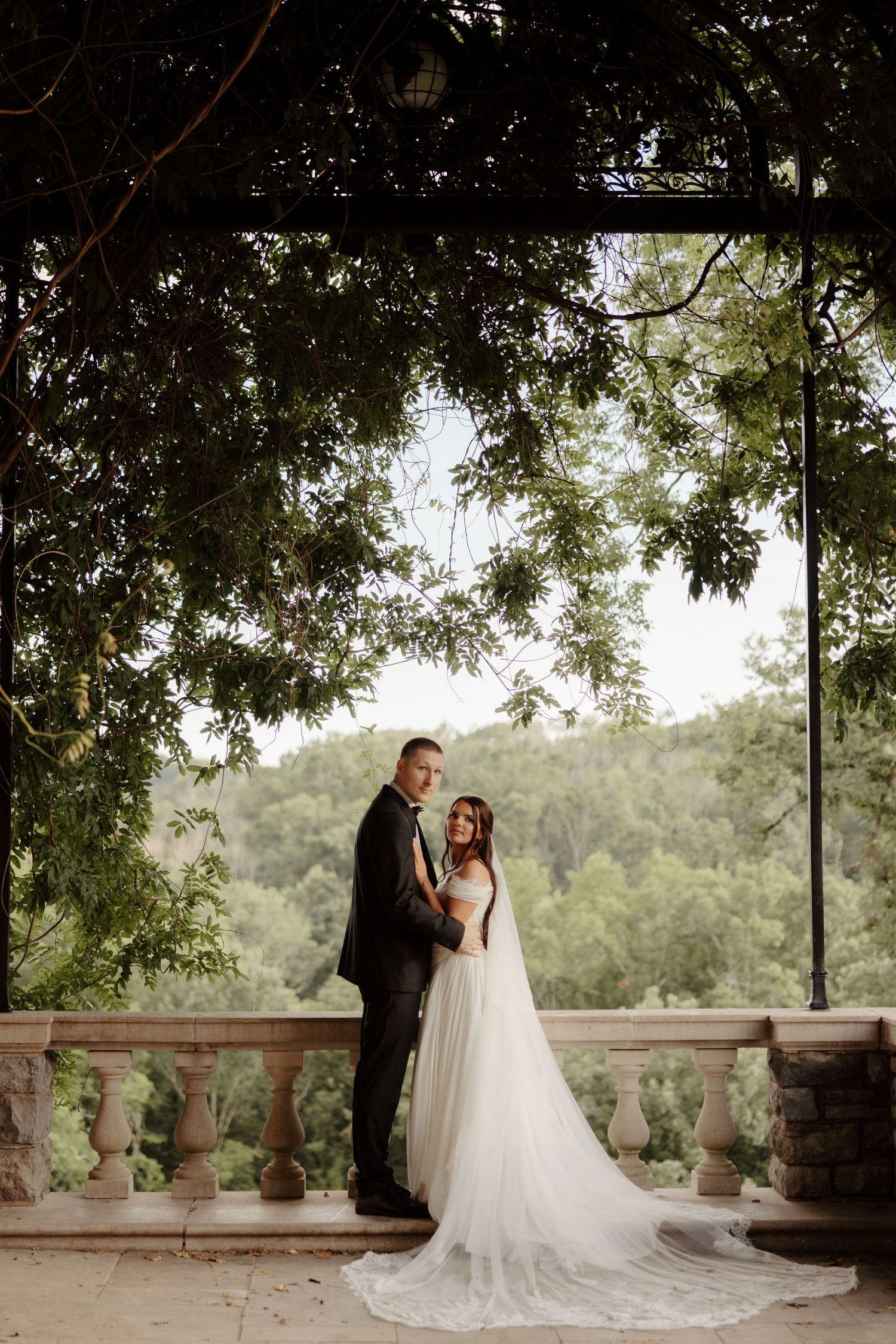 Bride and groom pose on stone balcony under lush green tree canopy with forested hills in background.