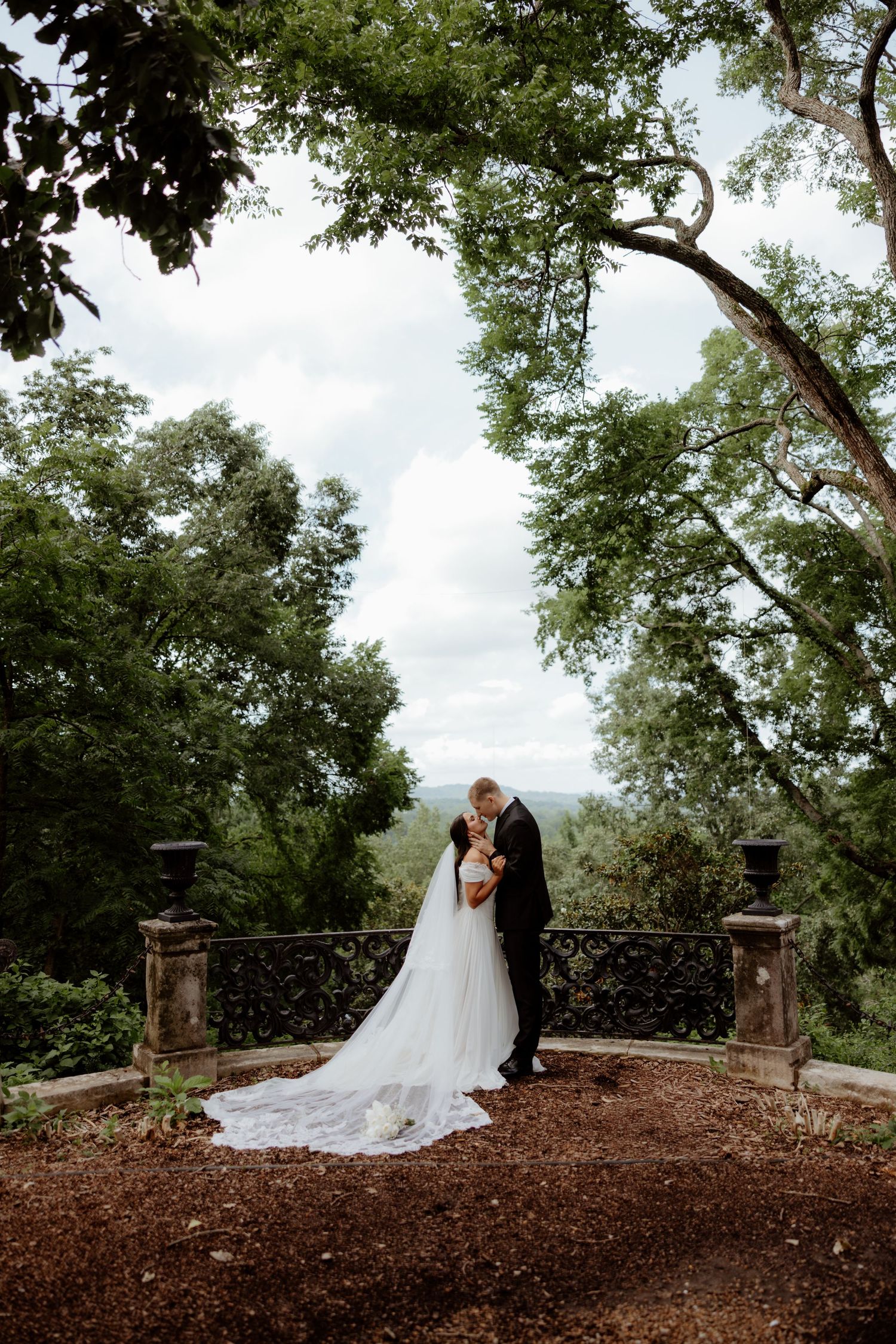 Bride and groom share a romantic kiss on a stone terrace surrounded by lush green trees on their wedding day.