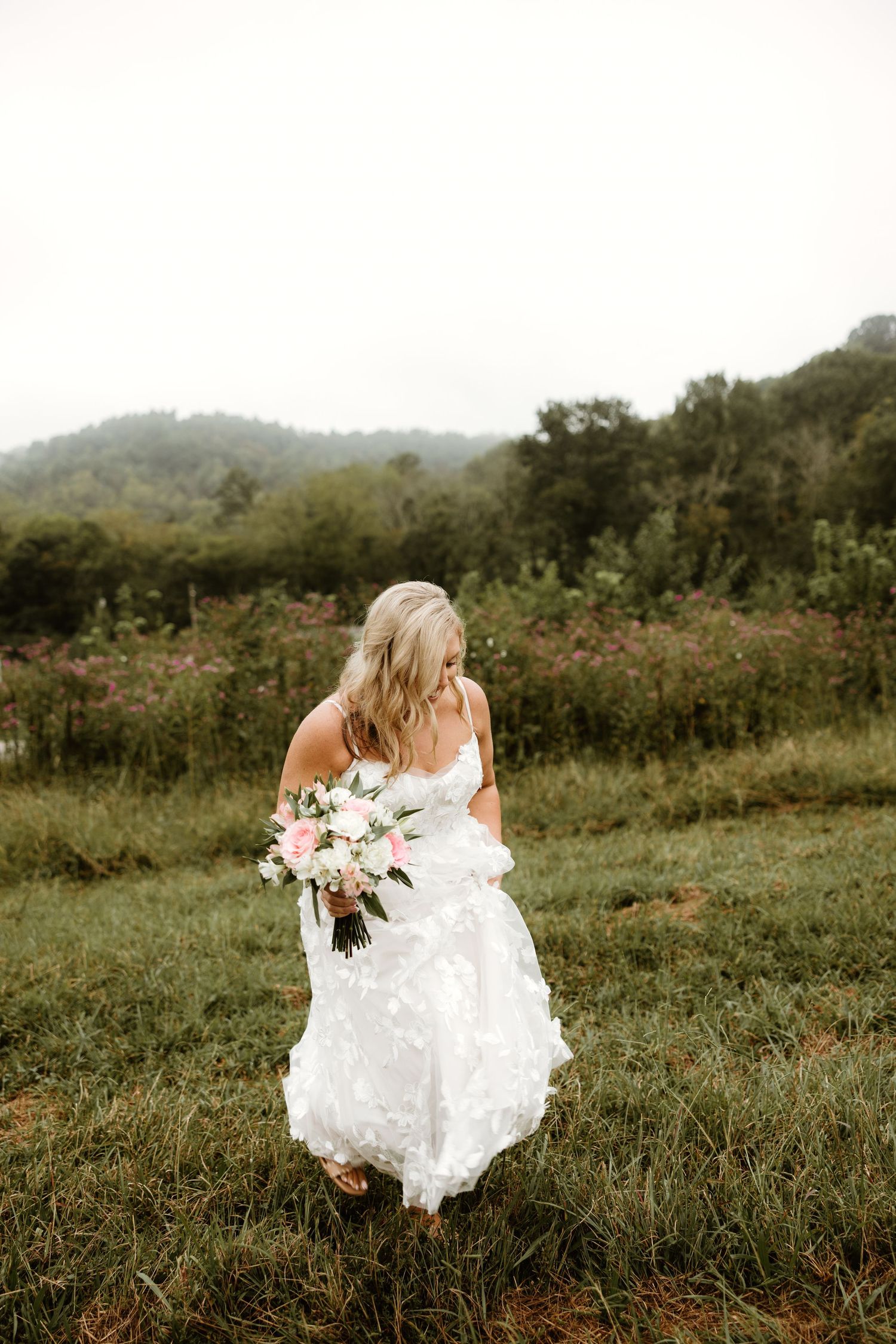 A bride in a flowing white dress runs through a misty mountain meadow holding a floral bouquet.