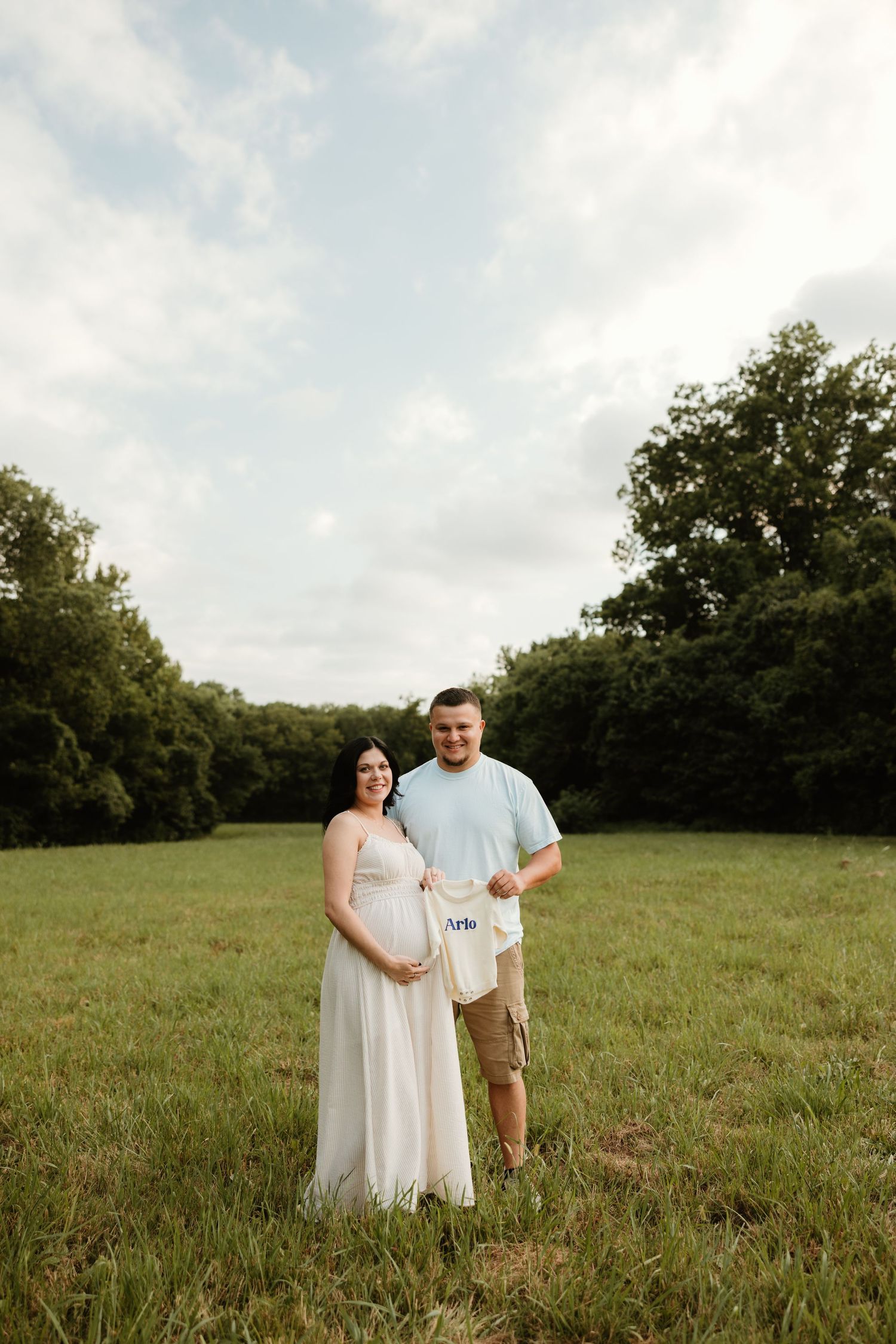 A couple in a grassy field poses together for a maternity photo, with trees in the background.