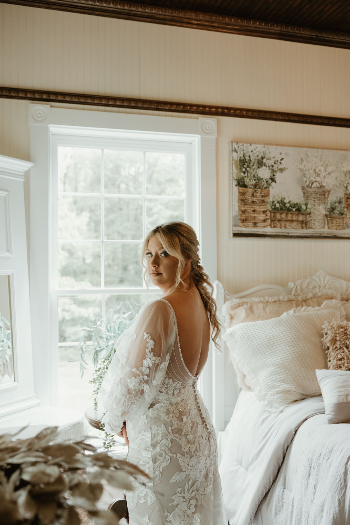 A wedding dress shows intricate lace detailing and an open back design in a sunlit bedroom.