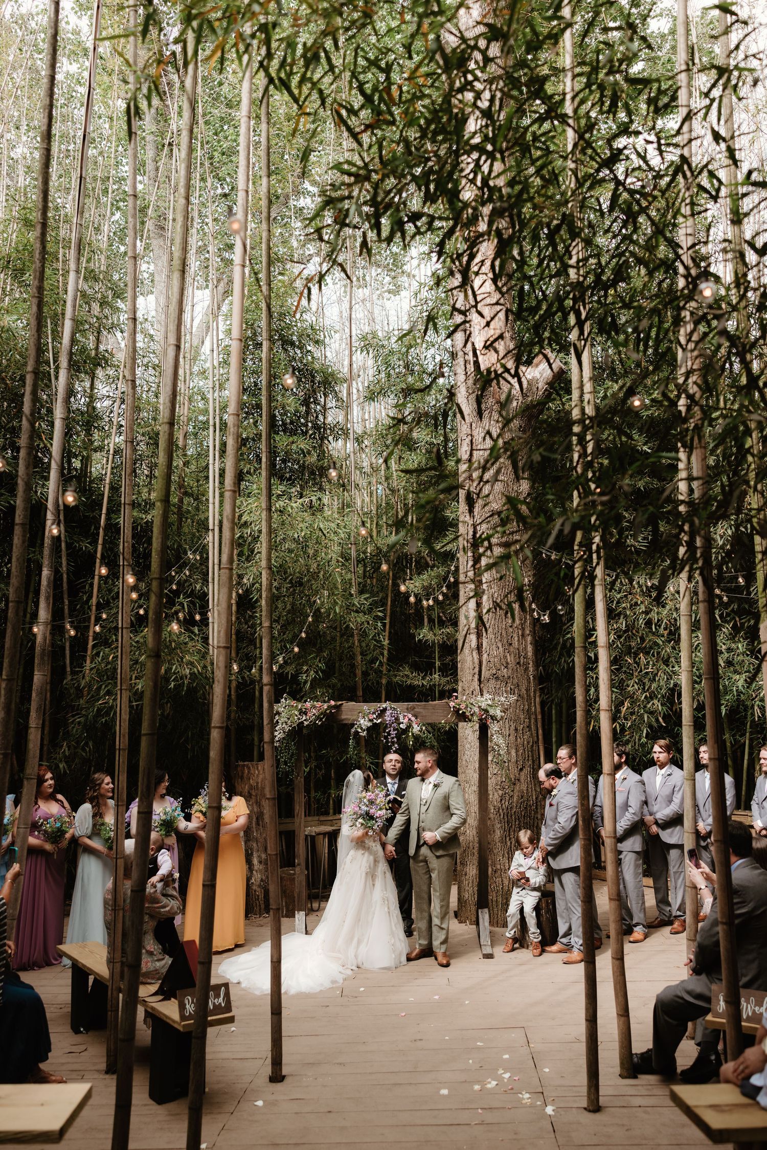 A woodland wedding ceremony takes place on an elevated wooden platform surrounded by tall trees and natural greenery.