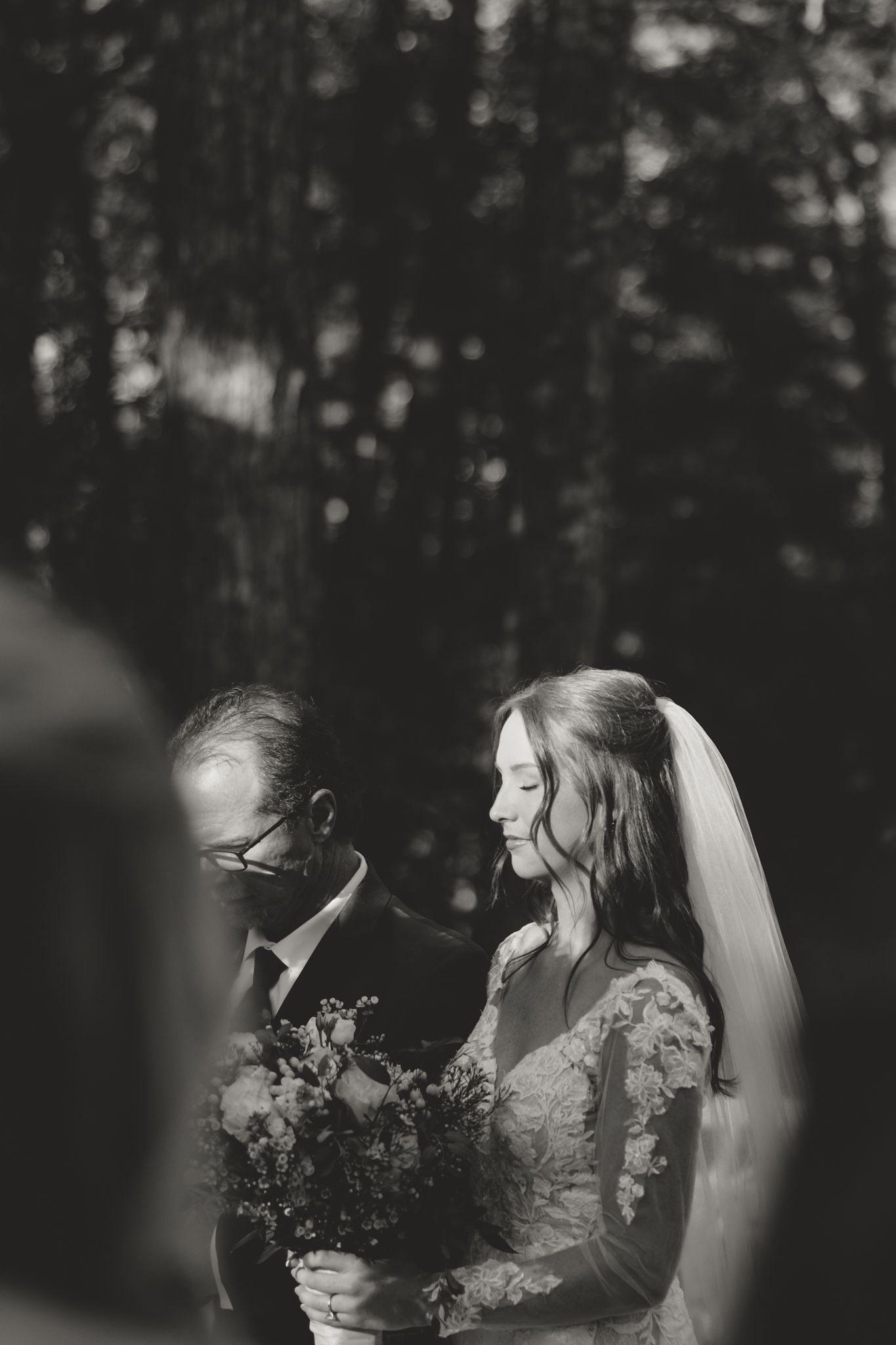 Black and white wedding photo with couple standing in front of wooded backdrop.