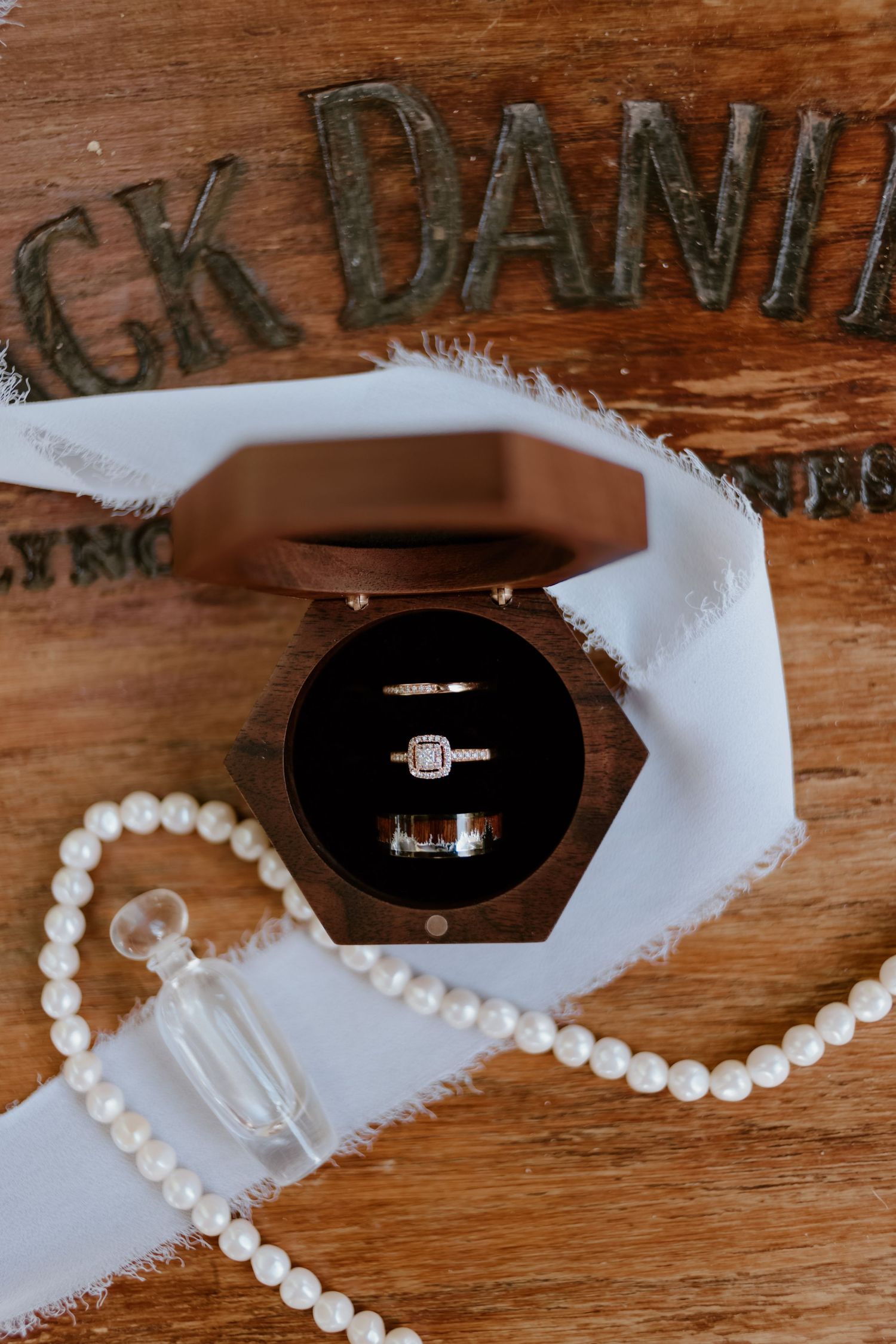 Wedding rings displayed in a wooden hexagonal box with pearl necklace and white lace on rustic wooden surface.