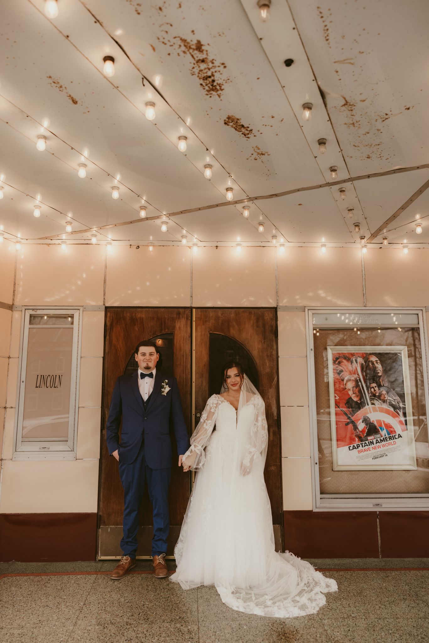 A couple stands beneath string lights in front of a vintage theater entrance on their wedding day.