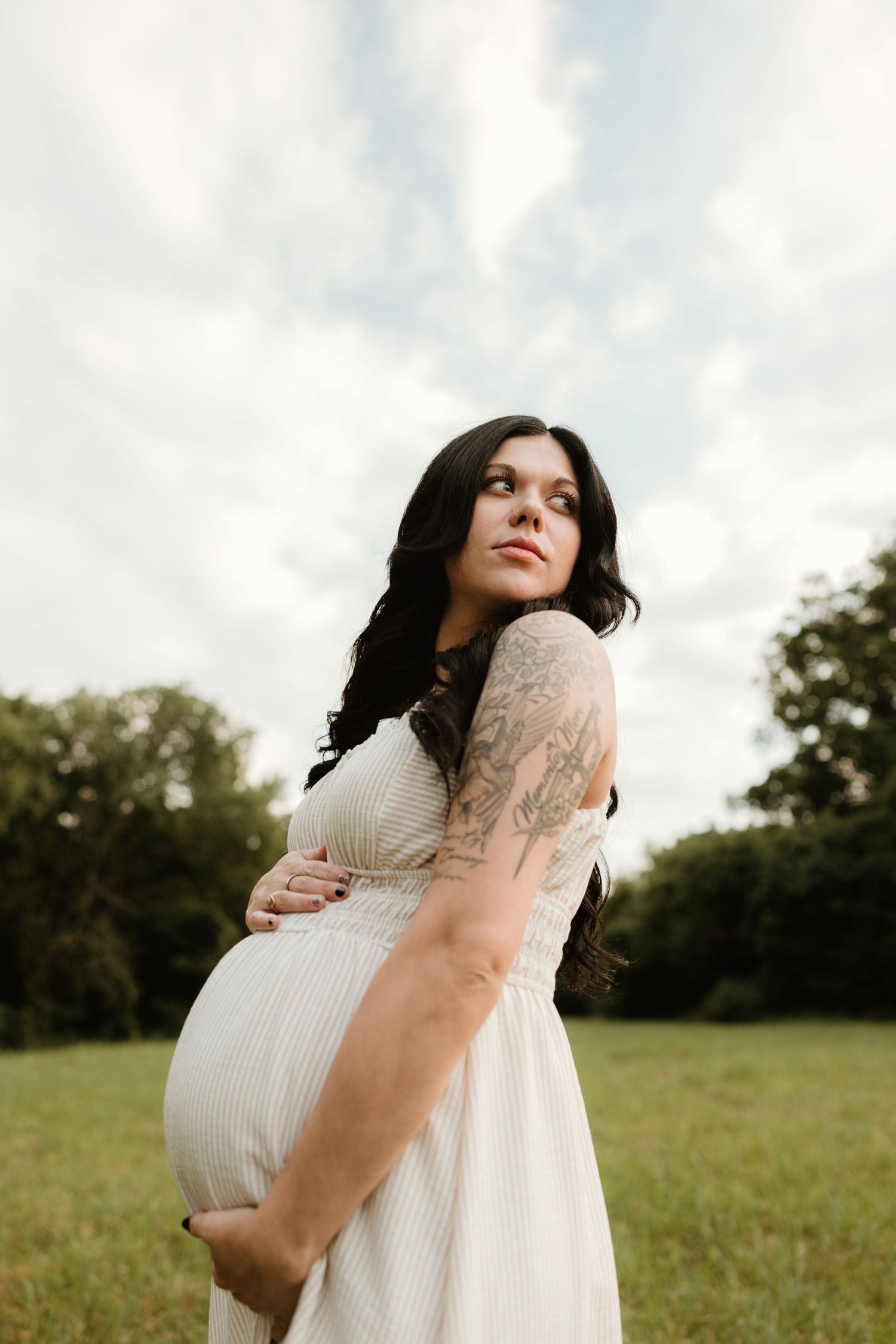 Expectant person in white dress stands in grassy field at sunset, gently cradling baby bump while looking upward.