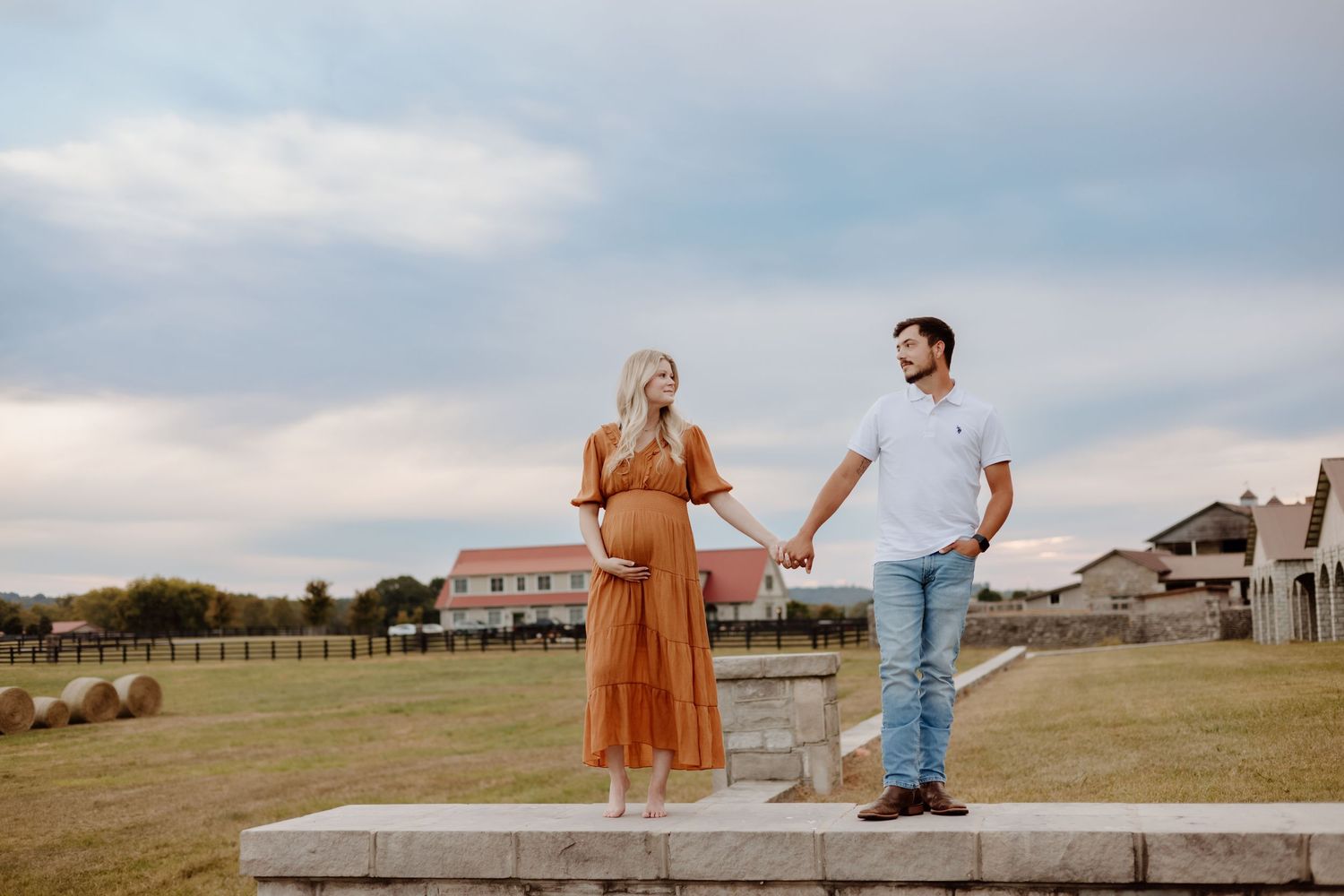 A couple holds hands while standing on a stone platform in front of a rural landscape with buildings in the distance.