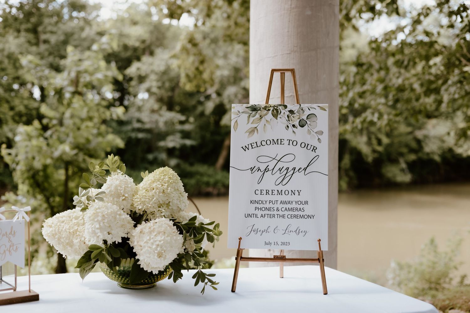 Wedding welcome sign displayed on gold easel next to white hydrangea floral arrangement at outdoor venue.