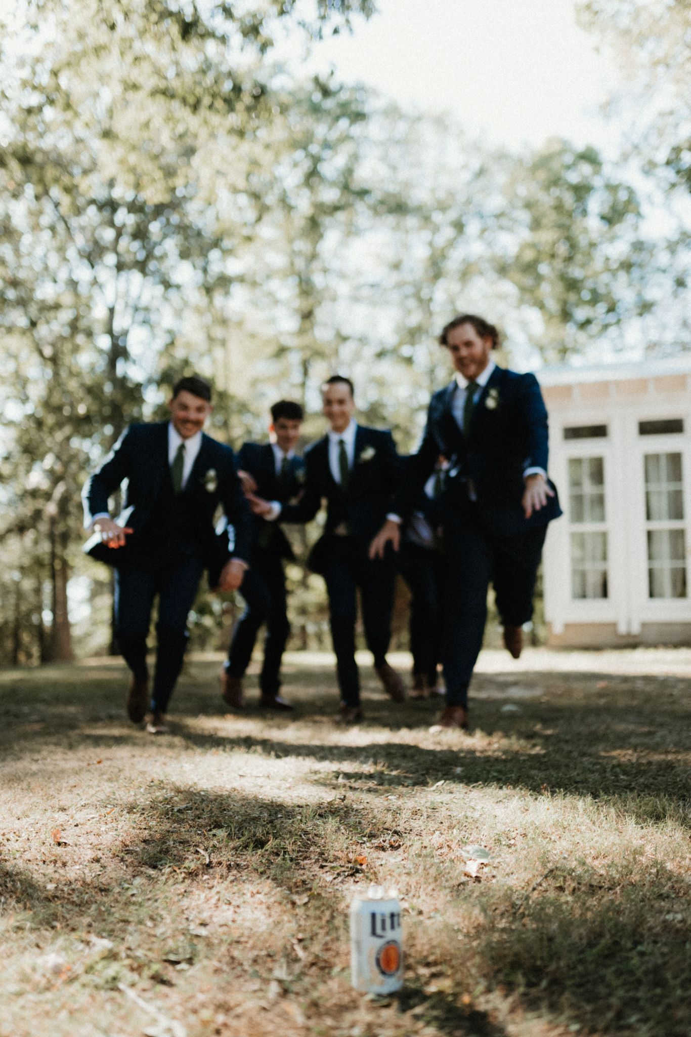 Groomsmen in dark suits run playfully across a grassy lawn during a wedding celebration.