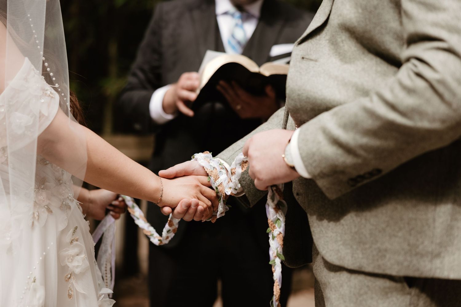 A couple holds hands during an outdoor wedding ceremony while an officiant reads from a book.