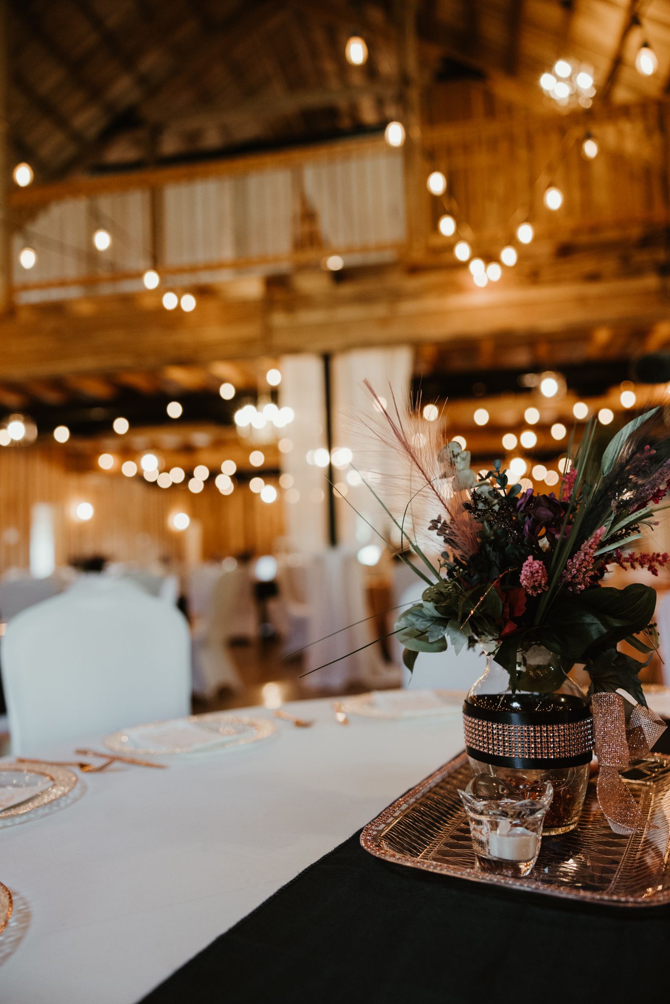 Rustic wedding reception table setup with string lights overhead and potted plants in copper vases on white tablecloths.