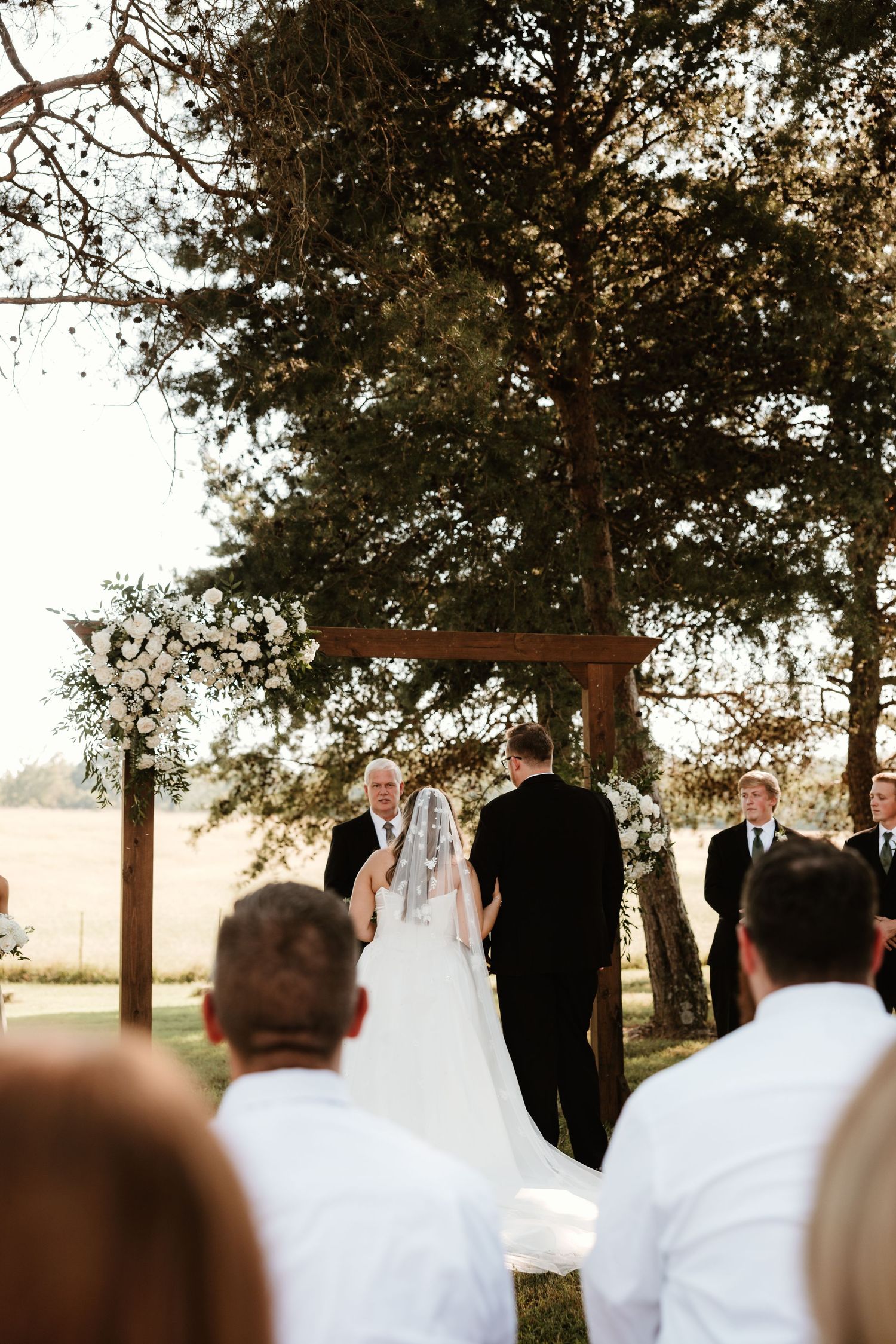 An outdoor wedding ceremony takes place under tall trees with a wooden cross arch at sunset.