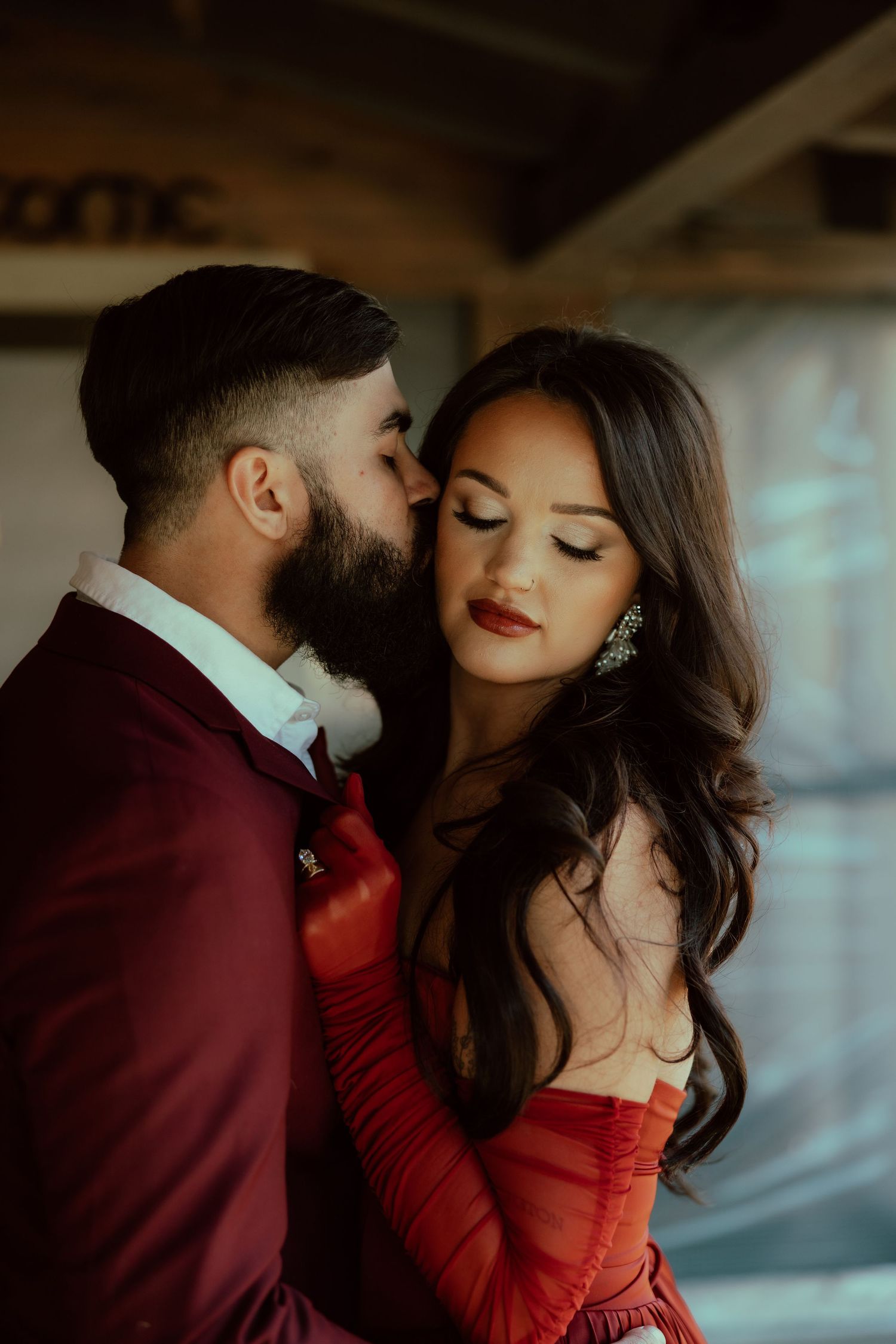 A bearded man in a burgundy suit kisses a woman in a red dress with dark wavy hair and bold red lips.